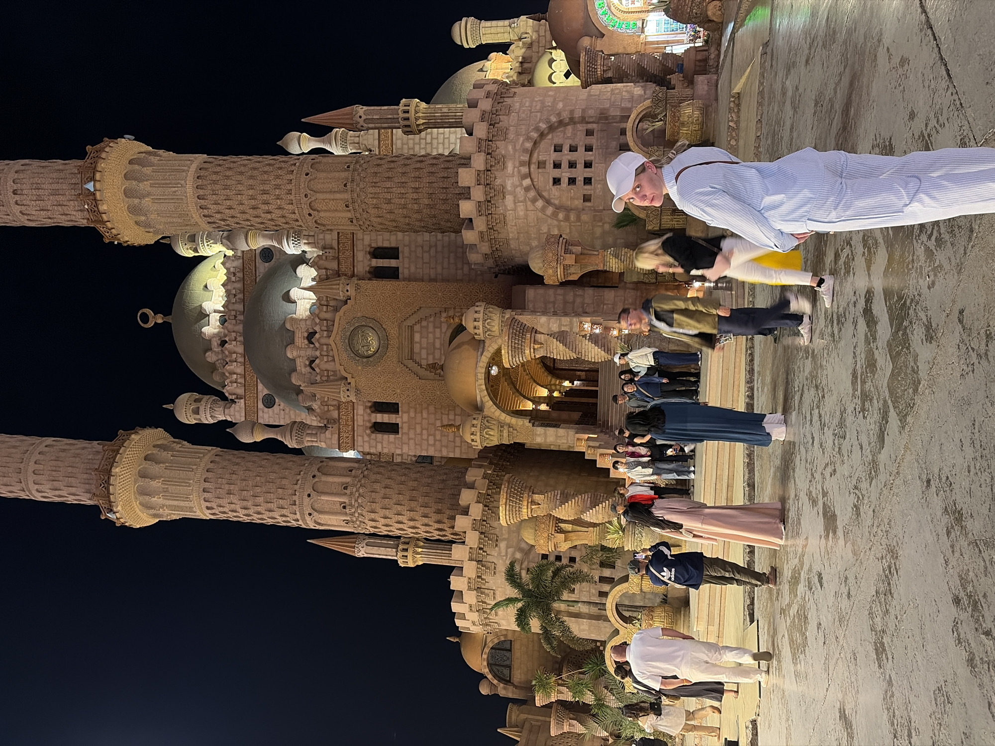 The image depicts the illuminated Al-Sahaba Mosque at night in Sharm El Sheikh, Egypt. The mosque, constructed from light-colored stone, features two prominent minarets with multiple balconies, several green domes, smaller spires, and intricate architectural details including arched entrances and Arabic calligraphy. Castle-like fortifications and battlements are integrated into its lower structure. The environment is an open, paved area of textured, light-colored concrete or stone. Approximately 20-30 individuals, predominantly adults, are present in the foreground and mid-ground.
In the immediate foreground, a woman wearing a light blue striped jumpsuit and a white baseball cap stands with her back partially to the camera, looking over her left shoulder towards the viewer. She has a dark strap diagonally across her back. Behind her, diverse individuals are scattered across the plaza. To the left, a man in a white polo shirt and white pants walks away from the camera. Towards the center, a person in a dark blue Adidas jacket photographs the mosque with a smartphone, while nearby, a woman in a long, light pink dress and another in a long, dark blue tunic stand near the mosque's steps. A larger group of people is gathered near the main entrance of the mosque. Palm trees and other sparse vegetation are visible near the base of the mosque on the left. Ambient lighting from the mosque and surrounding area illuminates the scene against a dark night sky. A partially visible, brightly lit green sign is present on the far right.