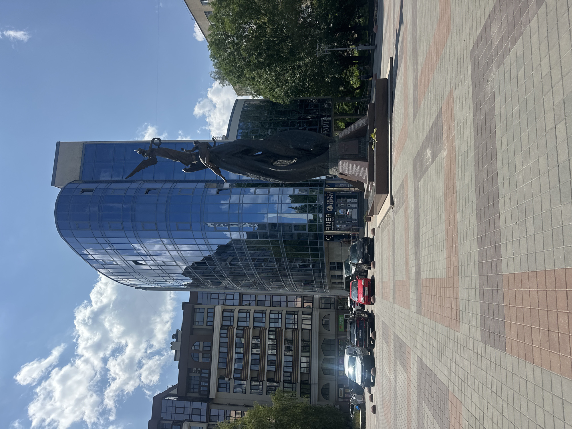The image depicts a street scene in Ternopil, Ukraine, featuring a modern glass-fronted building and a bronze monument in the foreground. The building, with its curved facade, reflects the blue sky and clouds. The monument, a multi-figured sculpture on a stone base, is prominently displayed in a paved square. Several cars are parked in front of the building, and a few trees line the right side of the scene. The sky is clear with scattered white clouds, indicating a sunny day. The pavement is made of interlocking rectangular stones in various shades of grey and red.