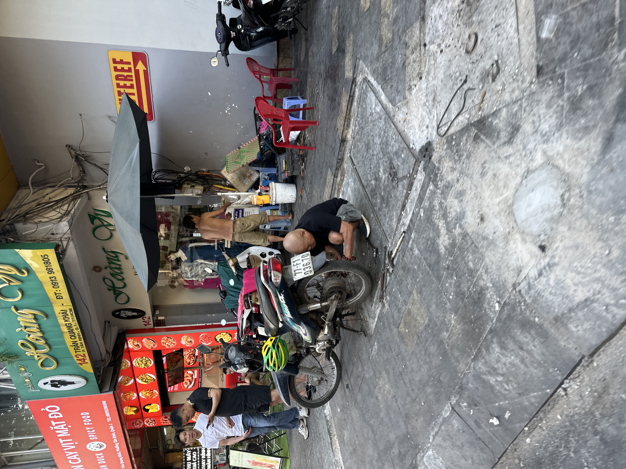 The image depicts a dynamic street-level commercial scene in Ho Chi Minh City, Vietnam. A visible shop sign identifies the location as 142 Trần Quang Khải, P. Tân Định, Q.1, TP.HCM.
In the foreground, a bald adult male, wearing a black t-shirt and grey shorts, crouches on the uneven concrete sidewalk. He is actively engaged in repairing the rear wheel of a dark blue/black Honda Wave Alpha motorcycle (license plate 77-F1 036.70), which rests on its kickstand. A green and yellow helmet is placed on the motorcycle's seat. Various tools, including wrenches and tire levers, are scattered on the ground near his work area.
To the left, two adult males stand conversing. One, wearing a white polo shirt, blue jeans, and white sneakers, holds a mobile phone in his left hand. The other, dressed in a black short-sleeved collared shirt with gold trim and black shorts, looks at his own mobile phone.
Further right and partially obscured by a dark grey open umbrella, a shirtless adult male wearing light brown shorts and sandals stands. He appears to be attending to a collection of items, including a white and a yellow bucket, a blue toolbox, and a green folding chair, near the entrance of a storefront. Two red plastic chairs and a blue plastic stool are also positioned in this area.
The storefronts include a "Hoàng Vũ" fashion shop (green sign) with mannequins in the window, and an adjacent "ẨM THỰC CAY VỊT MẶT ĐỎ" (Red-faced Duck Spicy Food) restaurant (red sign) displaying images of various dishes. A tangled network of electrical wires runs along the building facade above. On a grey wall to the right, a yellow and red sign with an arrow points right, featuring partial text "NTEREF." Several other motorcycles are parked along the sidewalk. The concrete sidewalk shows signs of wear, with cracks and small debris. The scene is illuminated by natural daylight.