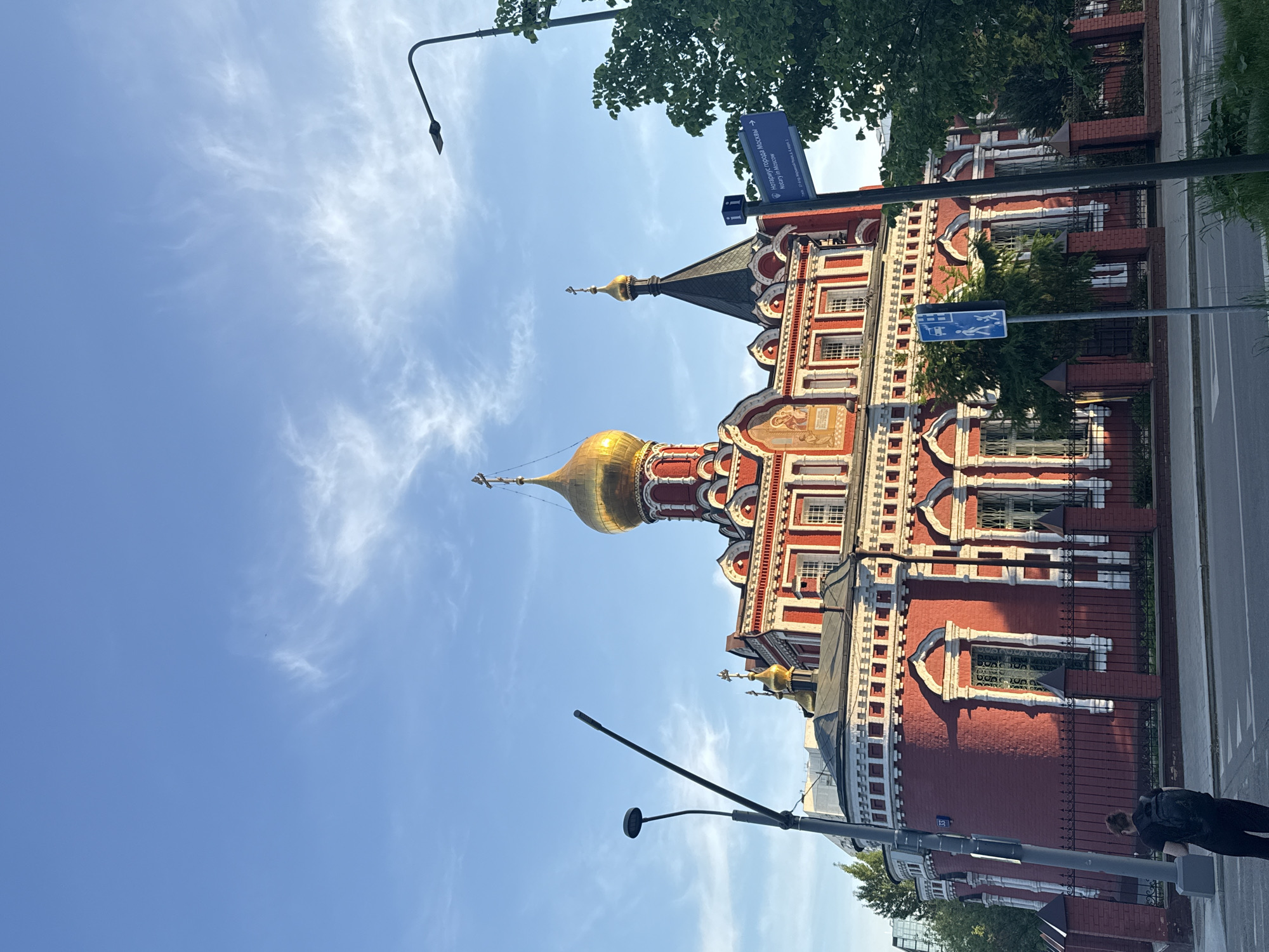 A red and white building with golden domes is seen in the city of Moscow, Russia. The building is next to a street with street signs and a streetlight. A person is visible walking near the building. The sky is blue with scattered white clouds.