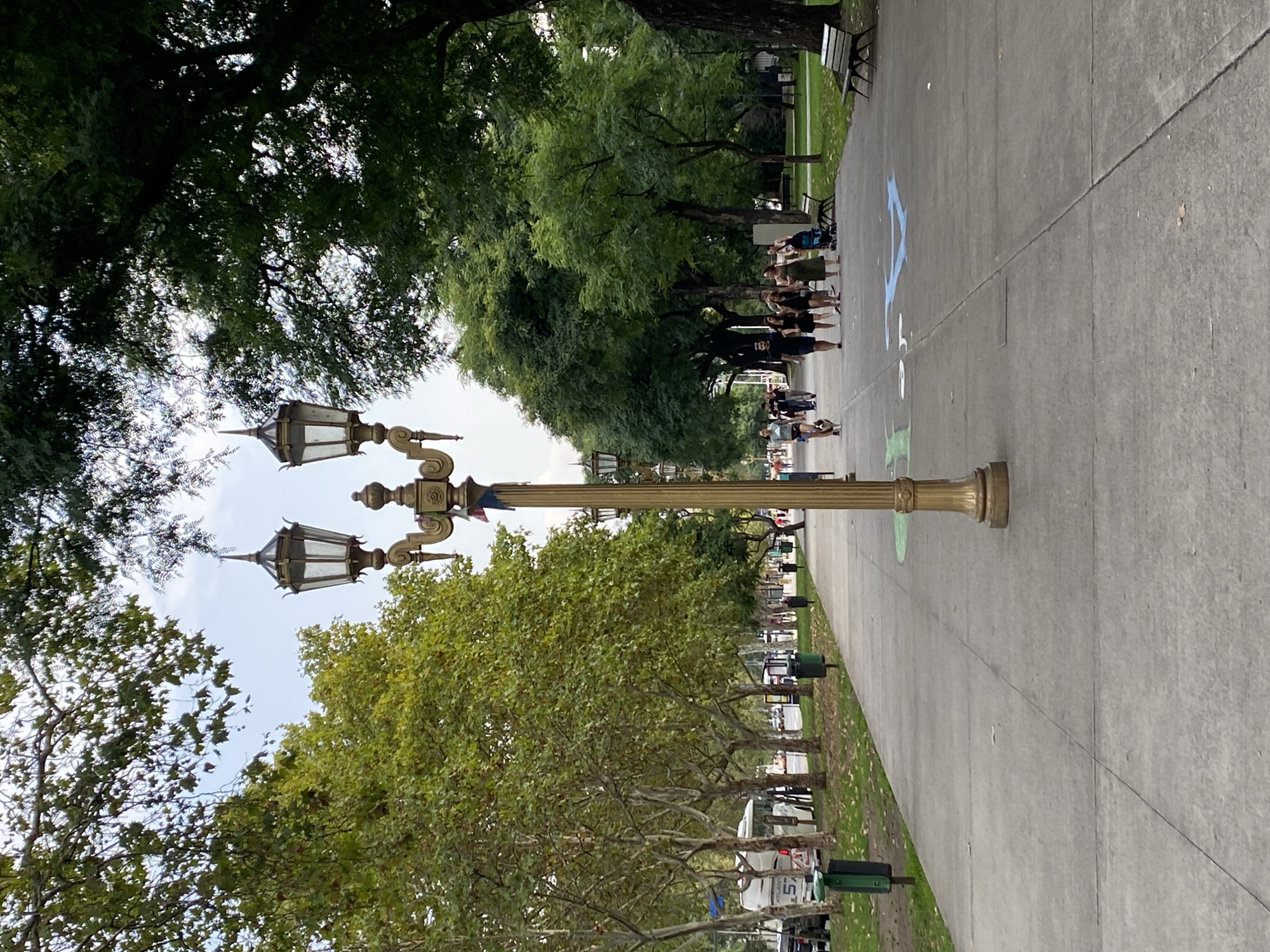 The image captures a scene in Buenos Aires, Argentina, along a wide concrete pathway lined with trees and vintage-style lampposts. A group of people stand in a row, partially obscured by trees, while others are further down the path. Chalk markings are visible on the pavement. Trees line both sides of the path, providing shade. Vehicles are parked along the street bordering one side of the park. Benches are visible along the path, and the sky is overcast.