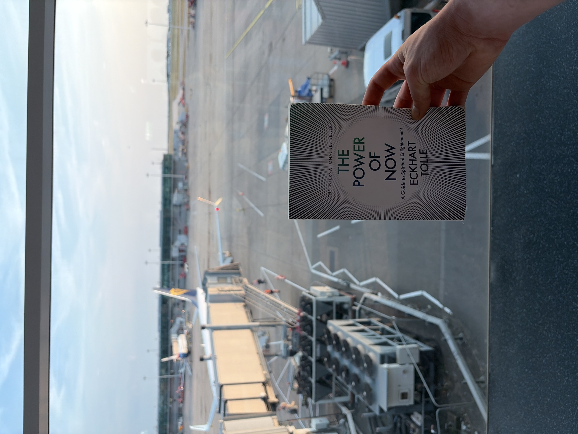 A person's hand is holding a book titled "The Power of Now" by Eckhart Tolle, subtitled "A Guide to Spiritual Enlightenment." The book cover features a stylized sunburst pattern and the text is predominantly green. The scene behind the hand and book is an airport tarmac, viewed from inside a building through a large window. Multiple airplanes, jet bridges, and ground support equipment are visible. The lighting suggests it is daytime, with the sky appearing bright and overcast. The overall impression is one of a waiting area or lounge at an airport, likely before a flight. The perspective is from above, looking down onto the tarmac.