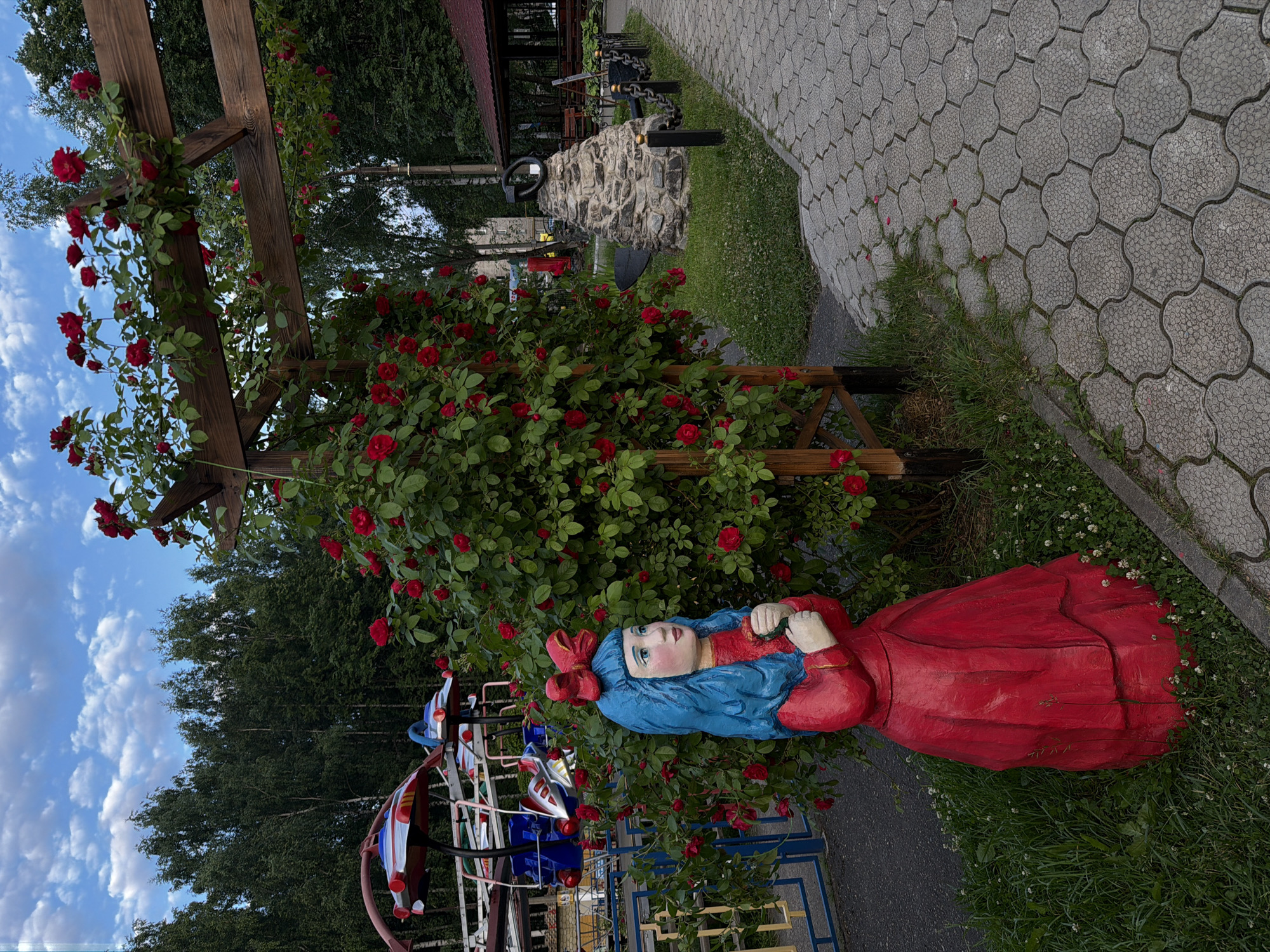 This media file shows an outdoor scene in Chaikovsky, Russia, featuring a wooden sculpture of a girl in a red dress with blue hair and a red bow. The sculpture is lying down amidst lush green grass and a vibrant bush of red roses. Behind the sculpture, a wooden pergola is covered with more red roses, some of which are in bloom. The scene also includes a paved pathway made of interlocking grey stones and a glimpse of a playground structure. In the background, a dense line of trees forms a backdrop, and a cloudy sky with blue patches suggests it might be late afternoon or early evening. The overall atmosphere is serene and somewhat whimsical, with the colorful sculpture and blooming roses adding a playful touch to the natural setting.