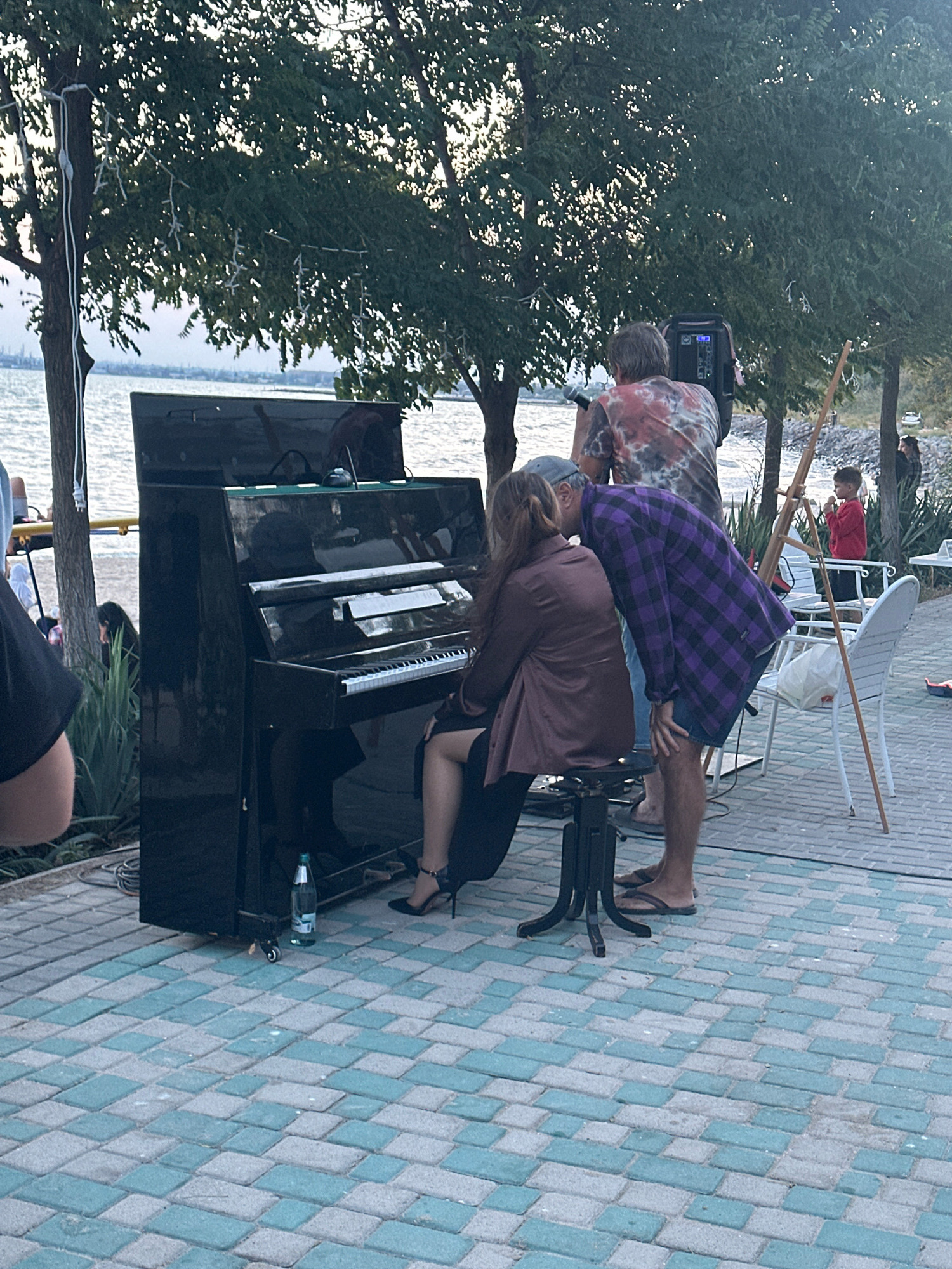 In Kryzhanivka, Ukraine, at a waterfront location, a woman is playing a black upright piano outdoors. She is seated and wearing a brown jacket and black skirt, with black high heels. Beside her, a person in a purple plaid shirt and denim shorts leans down towards the woman, appearing to be whispering or kissing her. Behind them, another person in a tie-dye shirt stands near a portable speaker, holding a microphone. The piano is set up on a paver stone patio with turquoise and gray tiles. The background shows a body of water, trees, and a few other individuals, including a child in a red shirt. A bottle of water is visible on the floor near the piano.