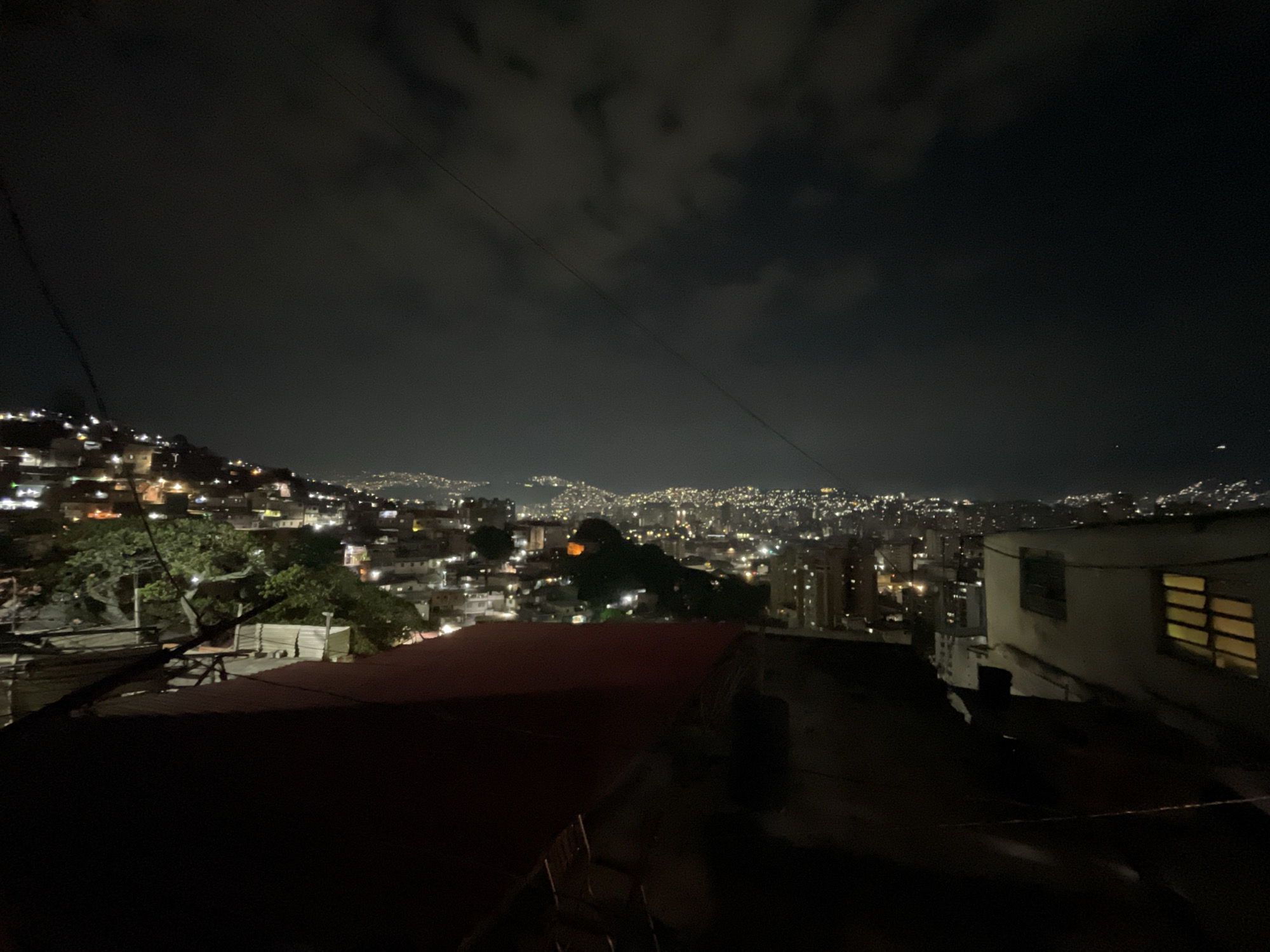 This is a nighttime cityscape view of Caracas, Venezuela. The foreground features rooftops, some with red surfaces, leading to a midground of densely populated hillsides dotted with lights. In the distance, city lights create a horizon glow under a cloudy night sky. There are no people visible. A single tree is partially visible, and some wires are strung overhead.