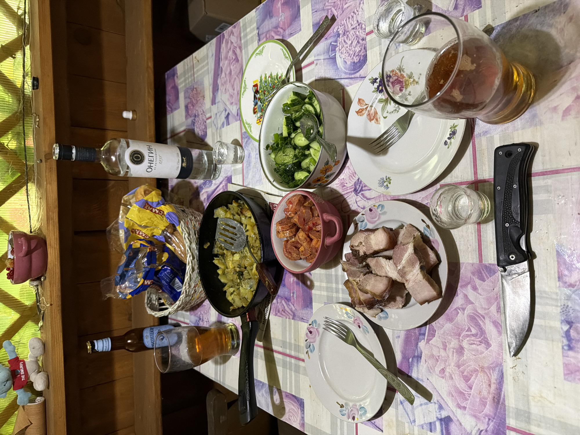 The image depicts a rustic wooden table set for a meal in what appears to be an outdoor or semi-outdoor setting, possibly a dacha in Medvedchikova, Russia. On the table, there is a bottle of vodka, a bottle of beer, a basket of snacks, a pan of cooked potatoes and vegetables, a bowl of chopped tomatoes, and a bowl of sliced cucumbers and dill salad. Two plates with slices of roasted pork are present, alongside cutlery and glasses. A knife is placed on the tablecloth. The background shows a wooden shelf with a stuffed toy and a pink bowl containing berries. The overall impression is of a casual, communal meal.
