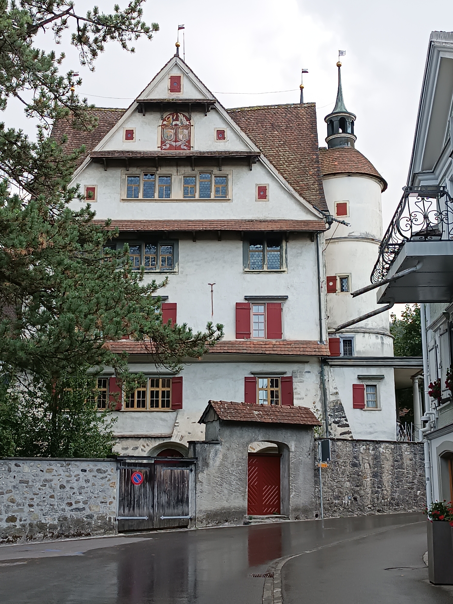 The image shows a traditional Swiss building with a white facade, red shutters, and a tiled roof. The building has a tower with a spire. A stone wall with a wooden gate is in the foreground. The street in front of the building is wet, suggesting it has recently rained. The building is located in the city of Appenzell, Switzerland, known for its picturesque architecture and rural charm. The overall impression is one of historical significance and architectural beauty.