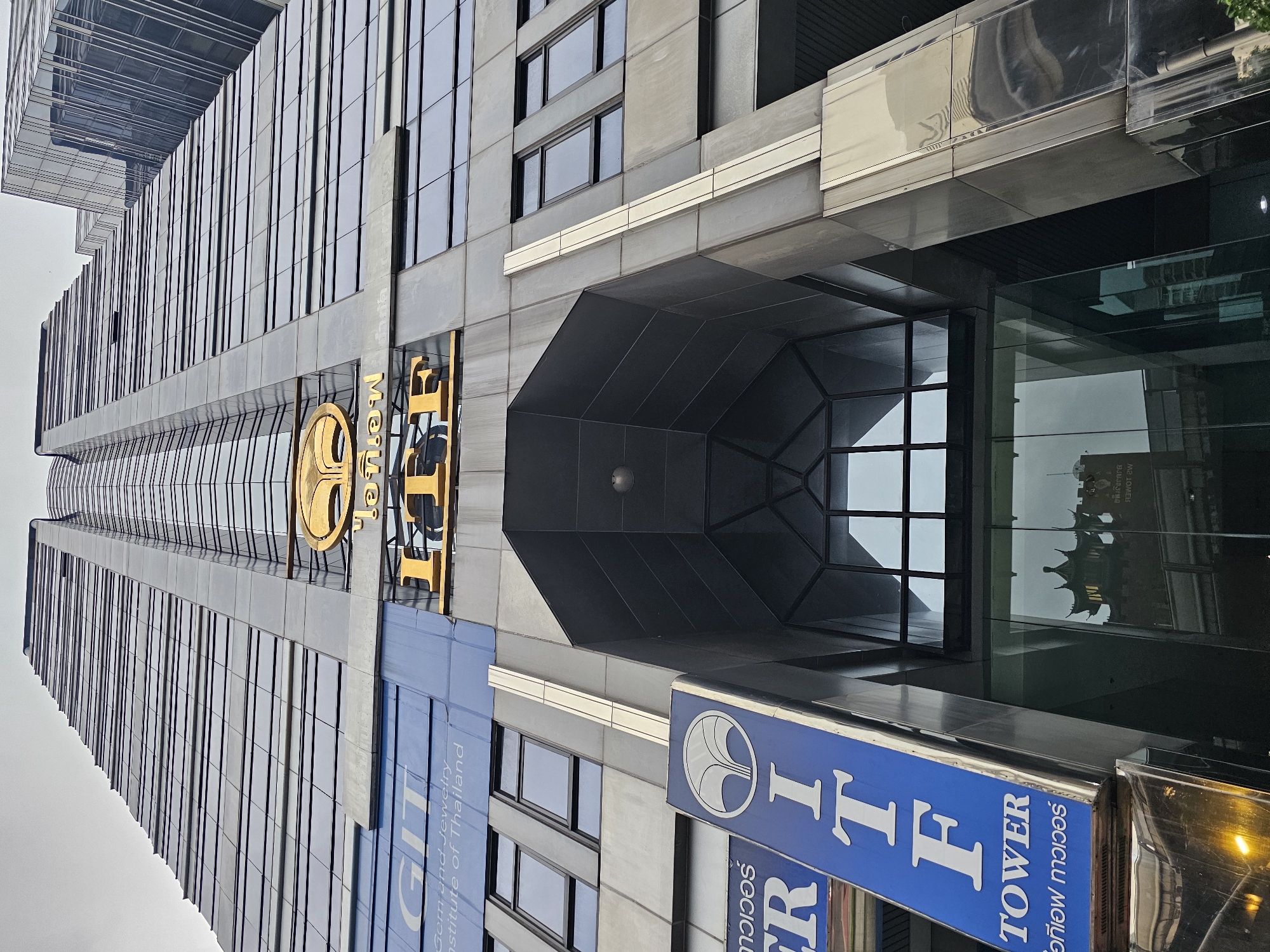 The image shows the ITF Tower, a tall skyscraper in Bangkok, Thailand. The building is made of glass and steel, with a distinctive golden logo at the top. The ITF Tower is located in the Silom district of Bangkok, a major commercial and financial hub. The area is characterized by a dense concentration of office buildings, hotels, and shopping malls. In the foreground, a blue sign with white lettering reads "ITF TOWER" and features a circular logo with a stylized tree. The reflection in the glass doors at the entrance of the building shows a traditional Thai temple, suggesting the presence of such structures in the vicinity. The sky is overcast, indicating a cloudy day.