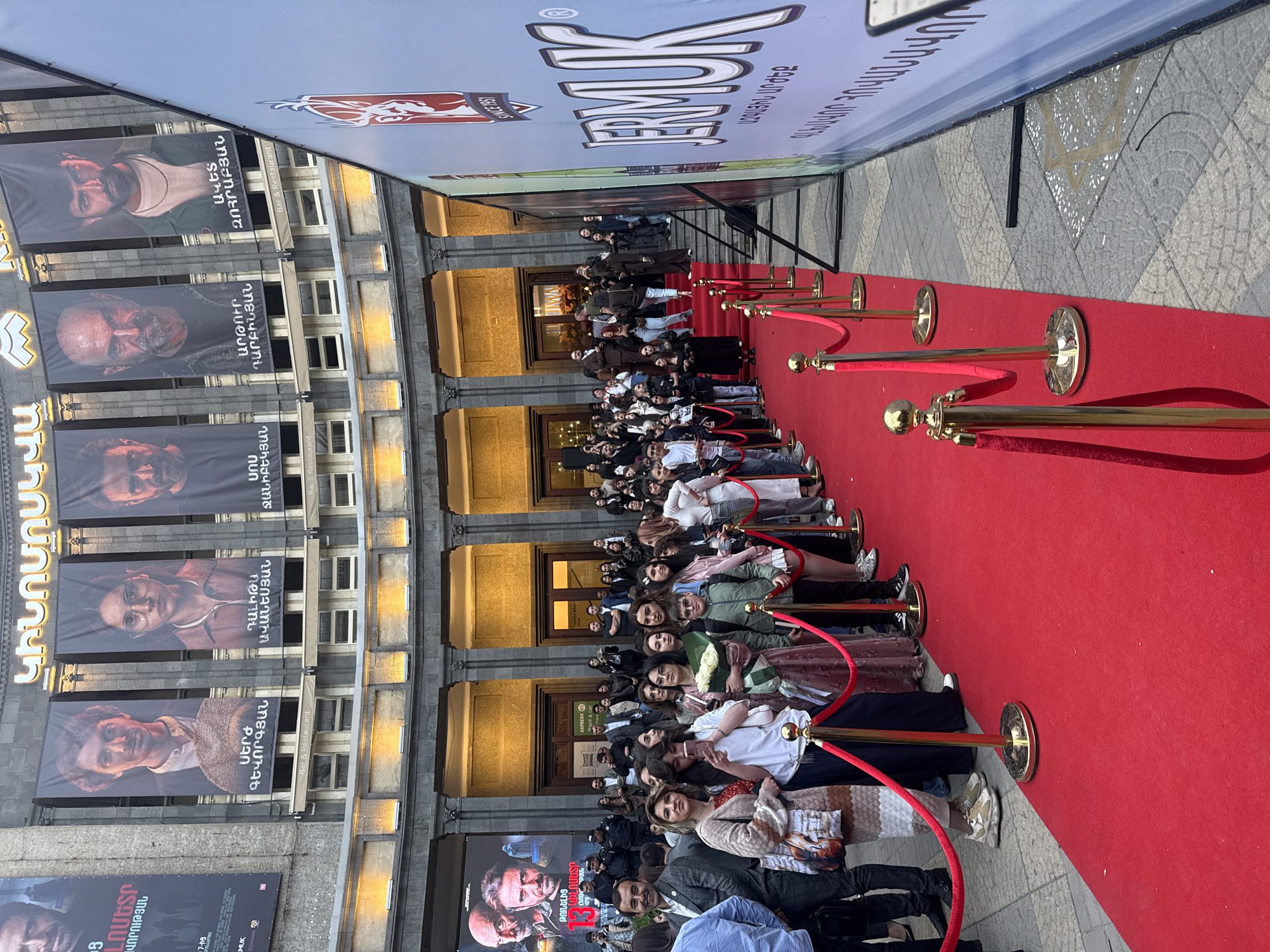 The image shows a crowd of people lining a red carpet leading to a building in Yerevan, Armenia. The building has multiple windows and is adorned with banners featuring images of people's faces and Armenian text. A large banner displaying the word "JERMUK" is visible in the background. The red carpet is lined with golden stanchions and red ropes. People are standing on both sides of the carpet, seemingly waiting for an event or premiere. Some are looking at the camera, and others are engaged in conversations. There is also text with the name "KINOMOSKVA" near one banner, suggesting that this is likely the Moscow Cinema.
