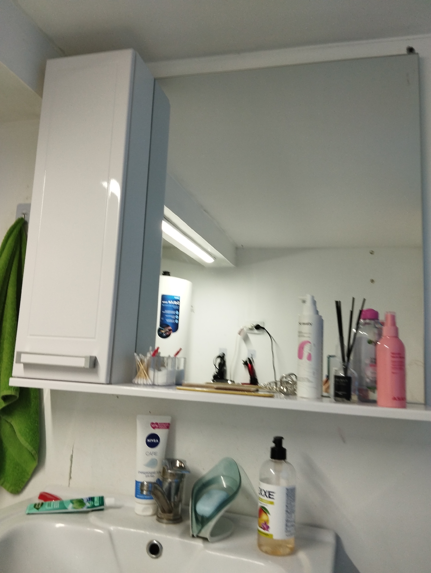 The image shows a bathroom scene, potentially in a residence. There is a white sink, with various toiletries arranged around it, including toothpaste, a tube of Nivea cream, a soap dispenser, and a washbasin. Above the sink, there's a shelf with additional items like cotton swabs, various bottles and containers of beauty products, and some decorative items. The shelf is mounted under a bathroom cabinet on the left and a mirror on the right. A green towel hangs on the left side of the cabinet. A fluorescent light is visible above the shelf in the mirror's reflection. Given the mention of Krasnogorskoye, Russia, the location could potentially be in that region, however, there are no elements in the image itself that pinpoint the exact geographic location.