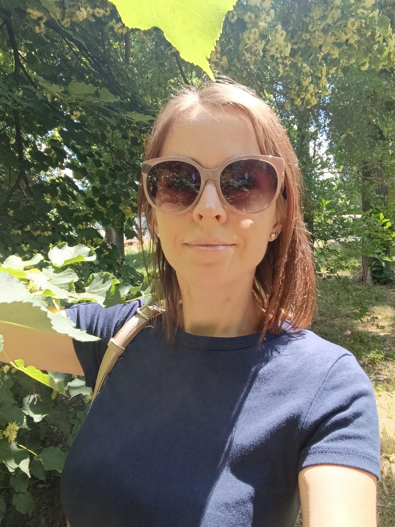The image is a close-up selfie of a woman with auburn hair, wearing sunglasses and a dark blue t-shirt. She is outdoors, with lush green foliage and trees in the background, suggesting a park or natural setting. The lighting indicates a sunny day. The woman is looking at the camera with a slight smile. The background shows trees and dry grass. Based on the provided information, the location is a park in Saratov, Russia.