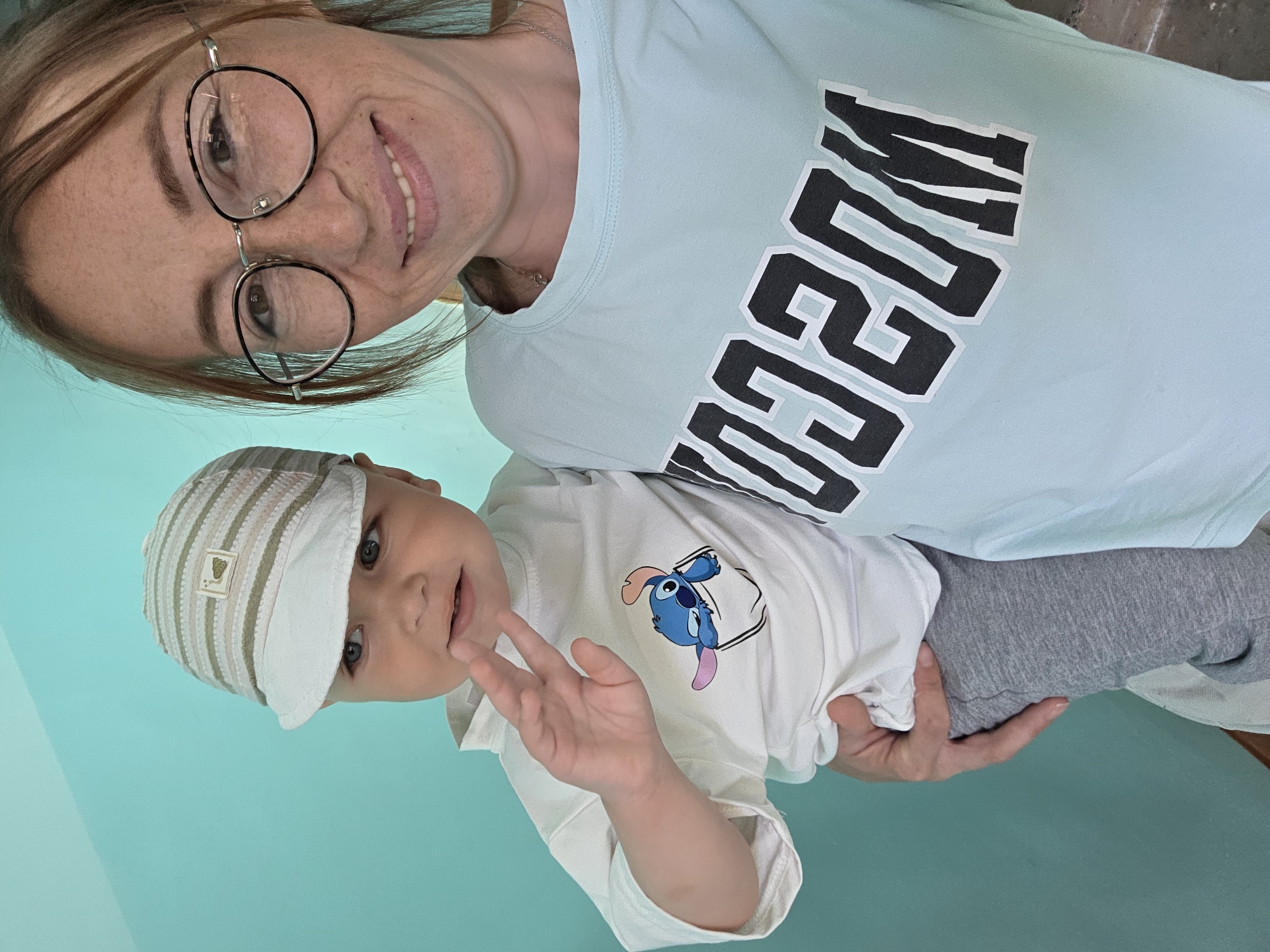 This is a selfie of a woman and a baby, taken indoors against a light blue wall. The woman, who appears to be in her late twenties or early thirties, is wearing glasses and a light blue t-shirt with "MOSCOW" in large black and white lettering across the front. She is smiling at the camera, and her hair is reddish-brown. The baby, held in her arms, is wearing a white t-shirt with a graphic of Stitch from Lilo & Stitch on it, and a striped cap. The baby is looking to the side and waving. The overall impression is a casual, happy moment. There are no visible elements that indicate a specific location within Chaikovsky, Russia, other than the likely residential setting.