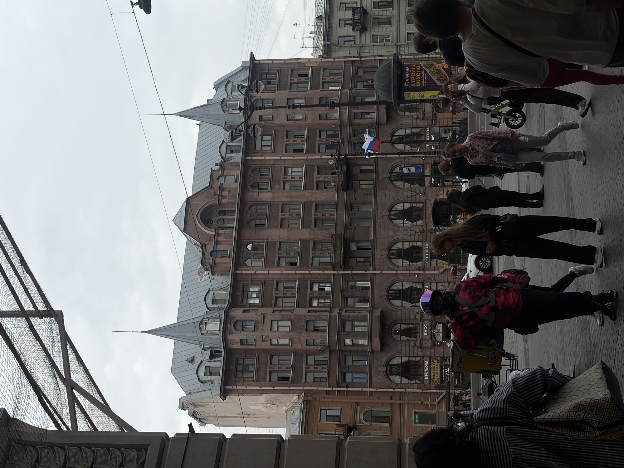 The image captures a street view in Saint Petersburg, Russia, on a cloudy day. A large, ornate building with reddish-brown facade and multiple windows dominates the background, featuring a distinctive mansard roof with spires and dormer windows. A Russian flag is visible flying from a lamppost in front of the building. In the foreground, several people are walking on a paved street. One individual, wearing a pink jacket and helmet, is carrying a yellow delivery box on their back. Other individuals are dressed casually in everyday clothing. Several businesses with signs in Russian are visible on the ground floor of the building. The overall scene suggests a typical urban environment in Russia.
