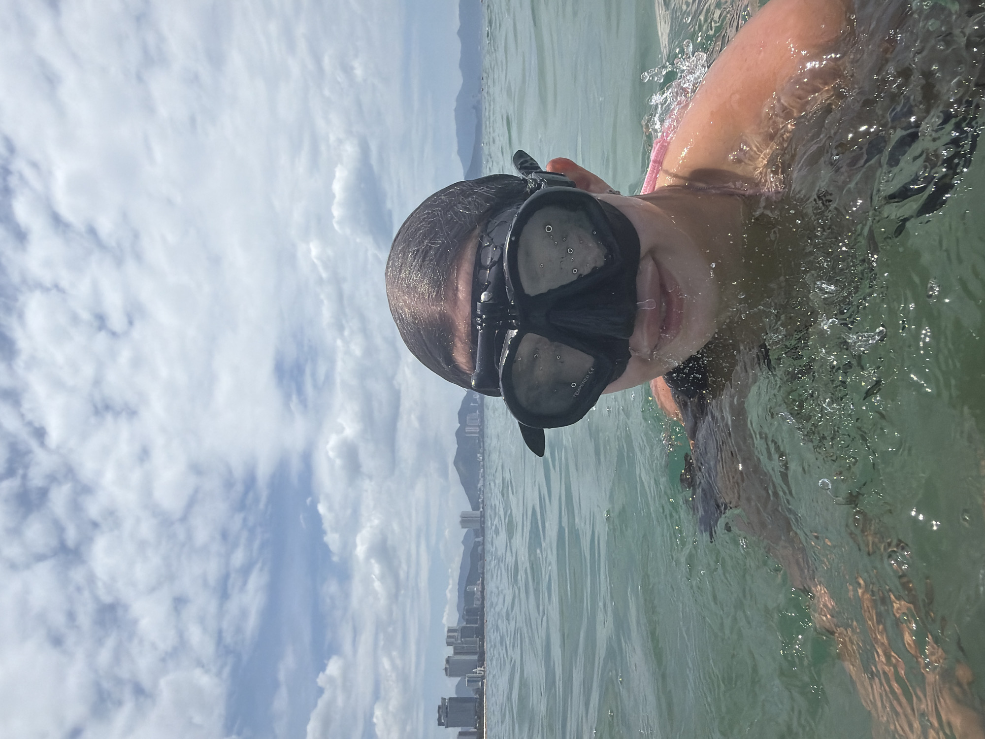 A person is swimming in the sea off the coast of Nha Trang, Vietnam, wearing a black diving mask and a pink strap bikini. The water is clear and green, with ripples and splashes around the person. In the background, a city skyline with tall buildings and mountains can be seen. The sky is cloudy and blue. The person's face is partially obscured by the mask, but a slight smile is visible. Water droplets are visible on the mask and on the person's chin. The person appears to be enjoying the water and the scenery.