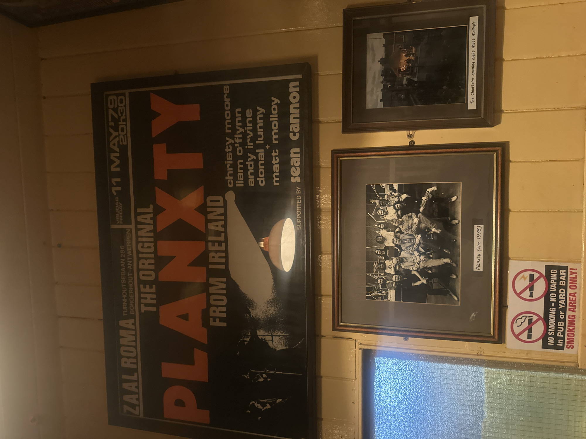 The image displays a wall decorated with framed posters and photographs, suggesting a venue related to music or performing arts. Dominating the left side is a large, dark poster for an event titled "PLANXTY - THE ORIGINAL FROM IRELAND." The poster features large, orange lettering and lists performers like Christy Moore, Liam O'Flynn, Andy Irvine, Donal Lunny, Matt Molloy, and Sean Cannon. Dates and location details are also visible: "FRIDAY 11 MAY '79 20h30" and "ZAAL ROMA TURNHOUTSEBAAN 286 BORGERHOUT-ANTWERPEN."
To the right, two framed photographs are hung. The top frame contains a black and white image with a handwritten label that reads "The Chieftains opening night Matt Molloy's." The bottom frame also holds a black and white photograph, captioned "Planxty (circ 1978)."
A "NO SMOKING - NO VAPING" sign with circular red and white prohibition symbols is prominently placed to the right of the framed photographs, indicating rules for the establishment. The overall setting appears to be indoors, with wooden paneled walls and tiled sections, possibly in a pub or bar, given the sign's text. The lighting suggests it might be evening, aligning with the concert poster's time. The atmosphere conveyed is one of historical and cultural significance, likely a place that celebrates Irish music and heritage.