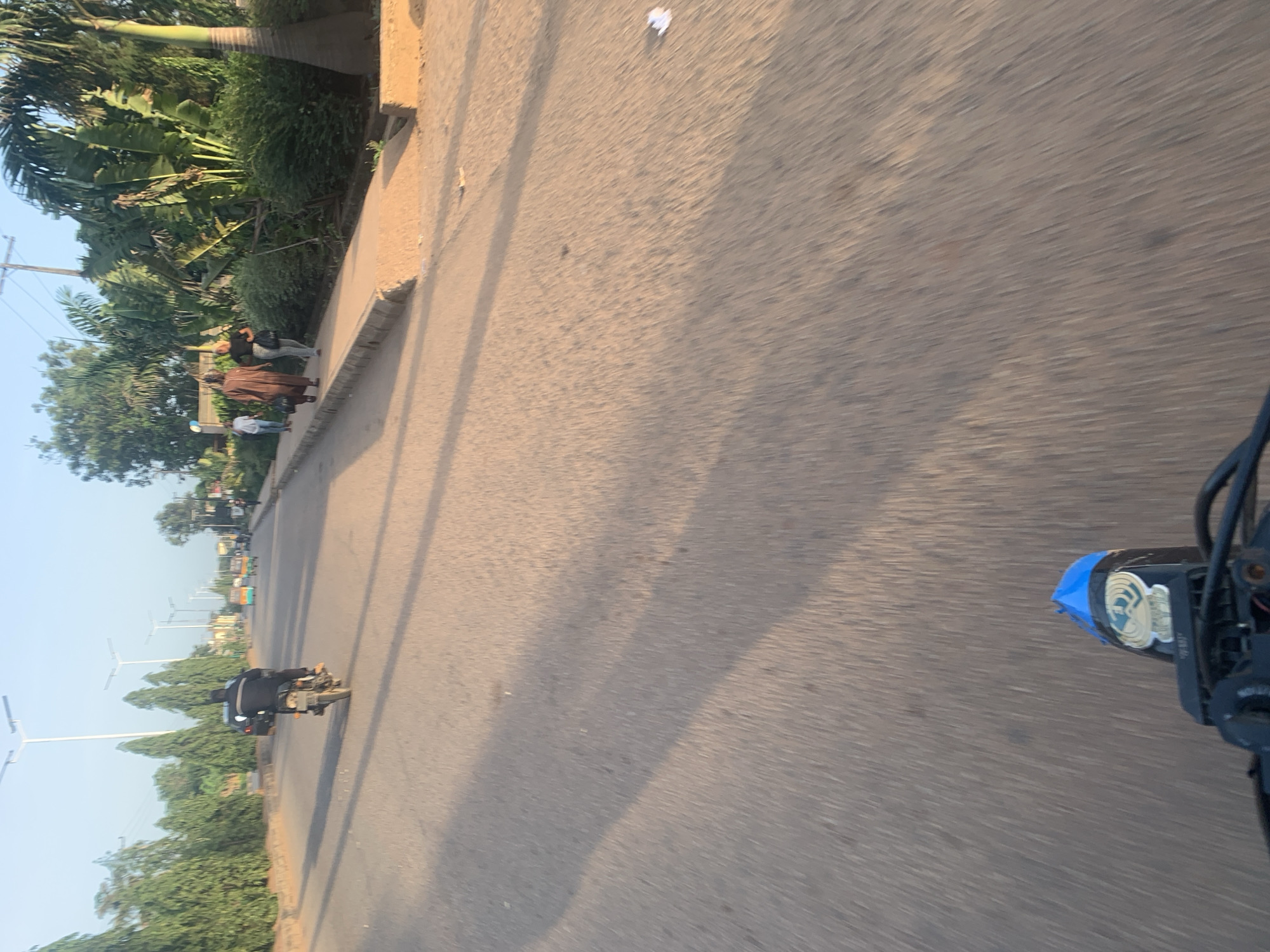 The image depicts a street scene in Jos, Nigeria, possibly captured during the late afternoon due to the long shadows. In the foreground, the handlebars and part of the headlight of a motorcycle are visible, indicating the photo is taken from the perspective of a rider. The road is paved with asphalt and shows signs of wear.
Further down the road, a person is riding a motorcycle, dressed in dark clothing. The motorcycle appears to be a standard commuter model. On the right side of the road, a paved sidewalk is present, with several individuals walking. One person is wearing a brown traditional garment, and another is dressed in casual attire. Behind them, dense green vegetation and palm trees line the area. On the left side of the road, a line of tall, slender trees creates a border. Streetlights with modern designs are visible along the road. In the background, there are more trees, some buildings, and other vehicles, including what appears to be a rickshaw-like vehicle. The sky is clear and light blue, suggesting fair weather. The overall impression is of a typical urban street in a developing country, with a mix of transportation, pedestrian activity, and natural elements.