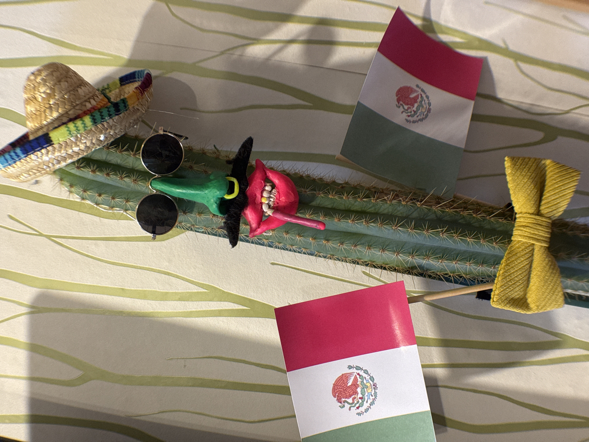 The image shows a cactus laid horizontally on a surface with a light-colored, patterned fabric. A miniature sombrero and a pair of sunglasses rest on the left end of the cactus. A green and red art sculpture is in the middle, with two small Mexican flags on either side. A yellow bow is located on the right end of the cactus. The location of Novo-Nikol'skoe, Russia, cannot be determined from the image.