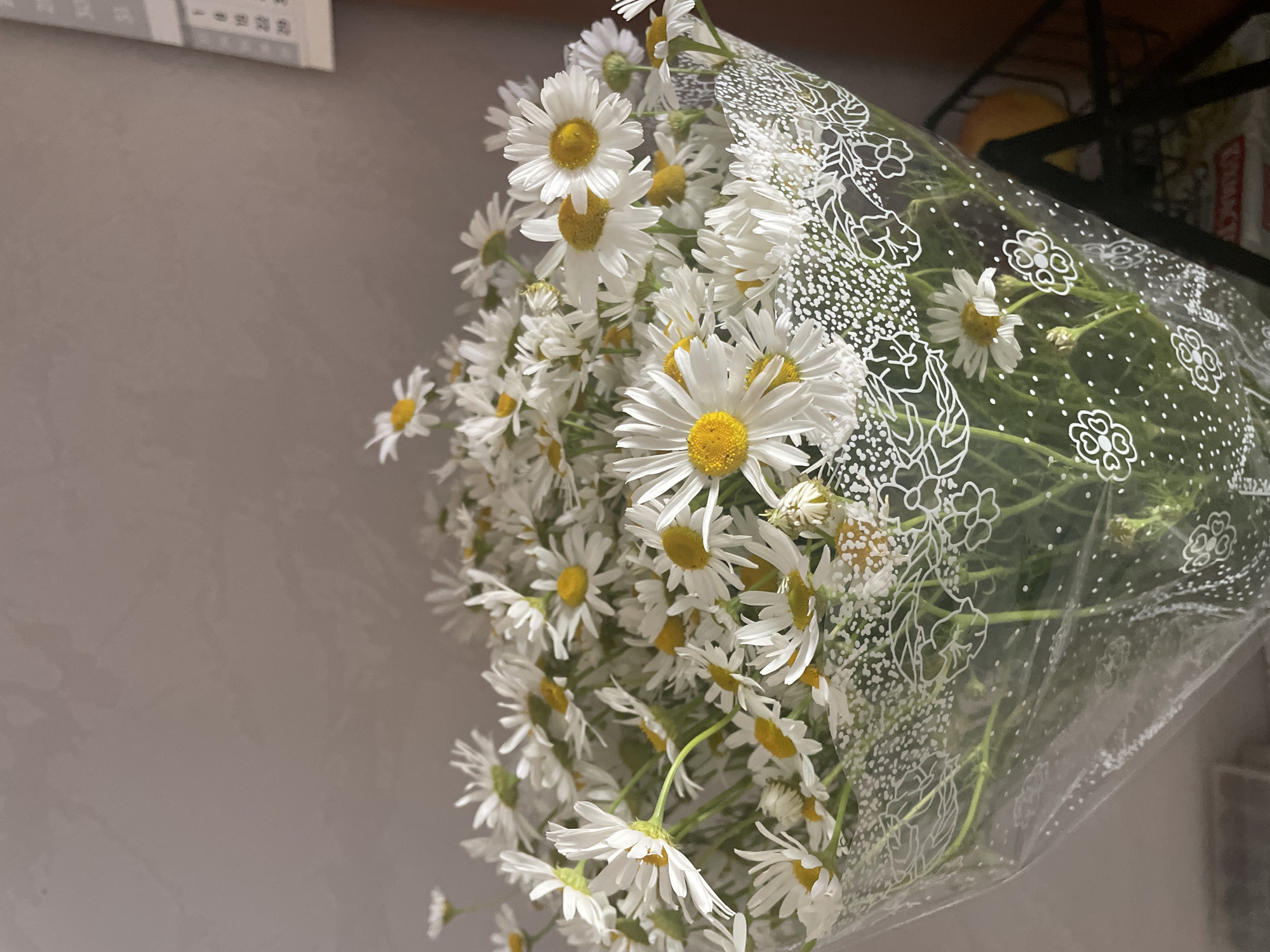 A bouquet of daisies is displayed in a clear plastic wrapper with a white floral design. The flowers are predominantly white with yellow centers, and their stems are visible through the wrapping. In the background, a neutral-colored wall is visible, with a calendar on the right side. Part of a dark metal shelf or rack is also visible in the lower right corner. The lighting suggests an indoor setting. The image is a close-up of the floral arrangement.