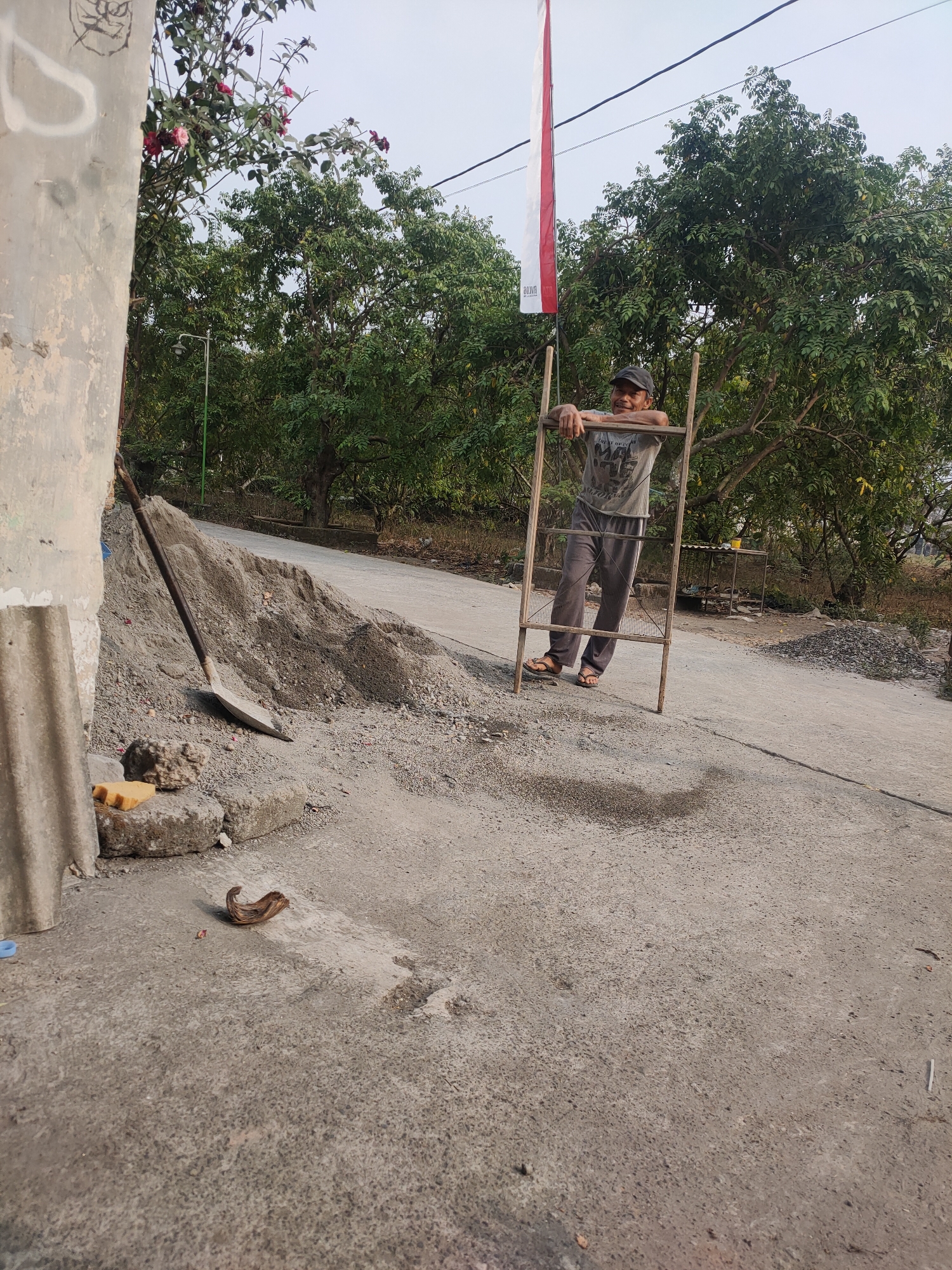 A man is leaning on a wooden frame on a dirt road in Kanigoro, Indonesia. The man is wearing a grey t-shirt, grey pants, and brown sandals. A red and white flag is visible behind him. In the foreground, there is a pile of sand and a shovel. A small snake is on the ground to the left of the frame. Trees and bushes surround the area.