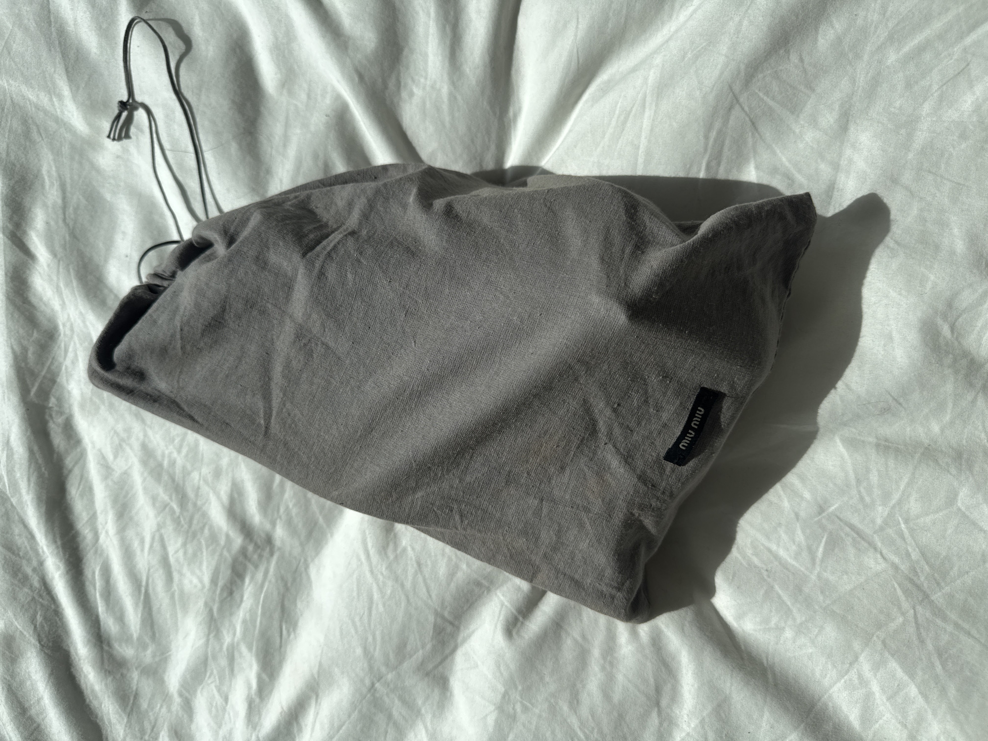 A gray drawstring bag with a black "miu miu" tag rests on a white, wrinkled surface. The bag is partially in shadow, and its drawstring is visible on the left side of the frame. The scene appears to be indoors, with natural light creating shadows on the surfaces. There is no indication of the location being in London, United Kingdom.