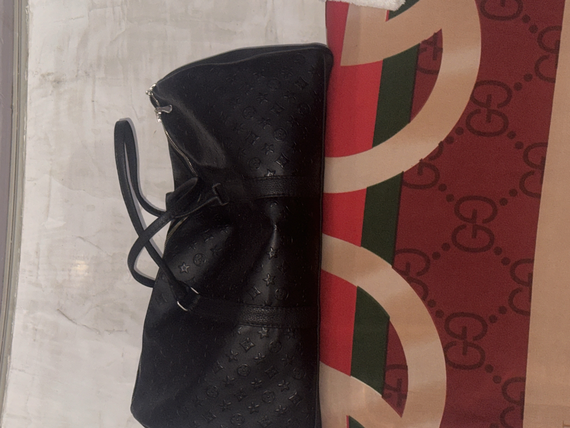 A black embossed duffel bag rests on top of a red and tan fabric with green stripes. The bag has two black handles and a zipper closure. The fabric beneath the bag features a repeating pattern of interlocking "G"s and dots. The background is a textured white wall.