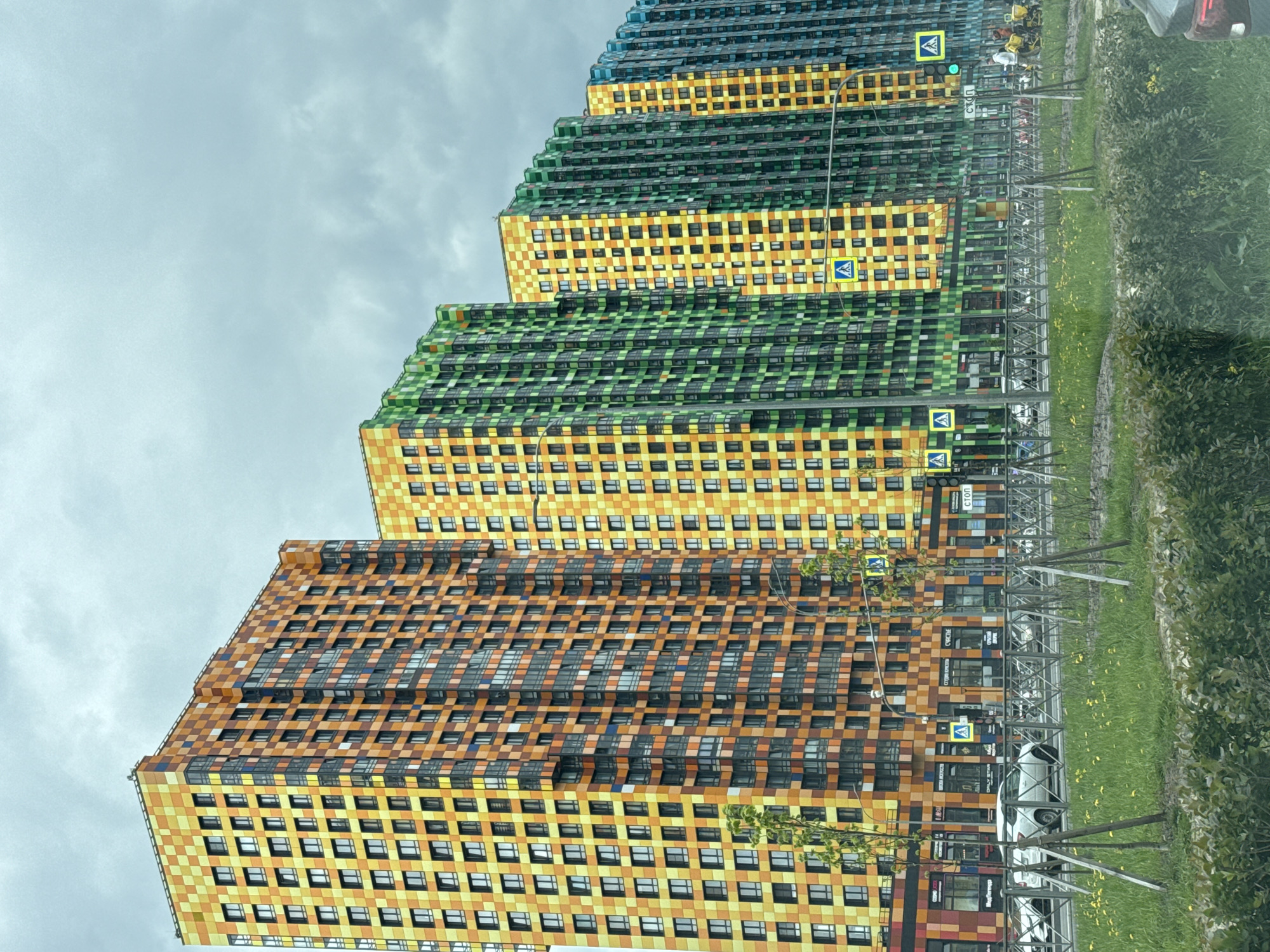 The image depicts several brightly colored apartment buildings located in Saint Petersburg, Russia. The buildings are designed in a step-like pattern, with each level progressively receding, and are colored in yellow, orange, green, and blue hues with a checkerboard pattern. Each building has numerous windows. In front of the buildings is a grassy area with small trees planted. There are pedestrian crossing signs and lampposts along the sidewalk. The sky is overcast. The image appears to have been taken from a moving vehicle.