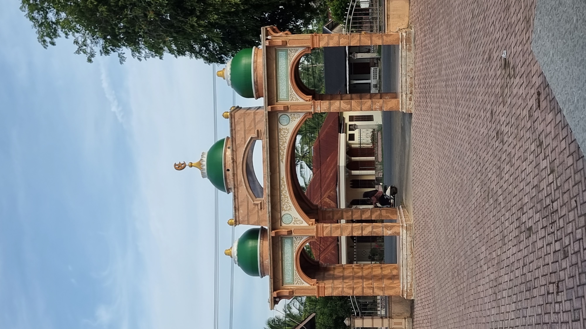 The image shows the entrance gate to a mosque in Meunasahlhok, Indonesia. The gate is constructed from light brown stone-like material and features two green domes topped with golden crescents. Intricate designs and Arabic script are visible on the archways. Behind the gate, a building with a red-tiled roof and white pillars is partially visible. A person is riding a white scooter through the archway. The ground in front of the gate is paved with reddish-brown interlocking bricks. The sky is a clear blue with some scattered clouds. A large tree with green foliage is visible on the right side of the frame. A metal fence runs along the sides of the property.