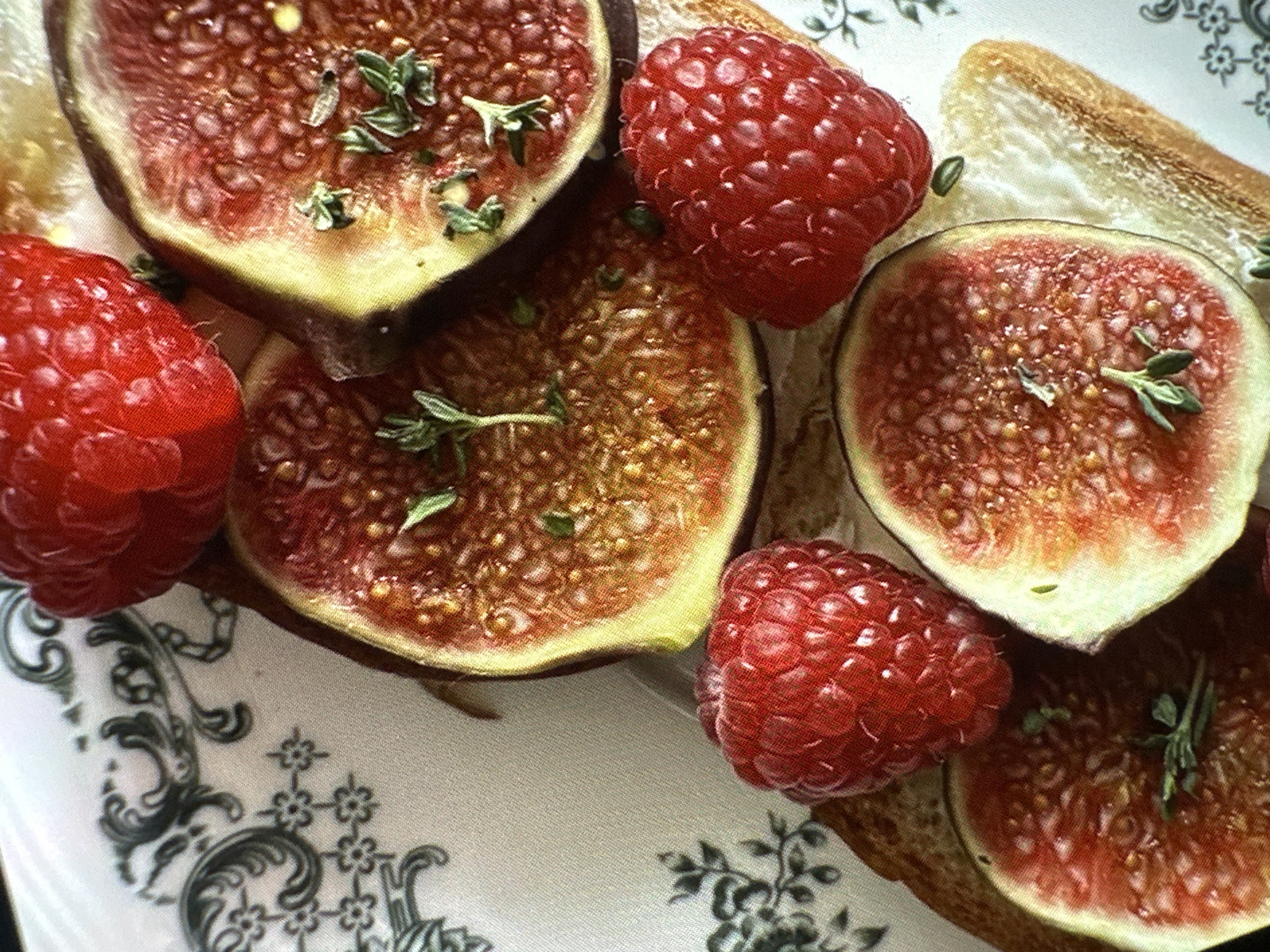 A close-up shot presents a plate of food, likely a dessert or appetizer, featuring figs and raspberries on what appears to be toast. The figs are sliced in half and topped with sprigs of green herbs, possibly thyme. The raspberries are plump and red, scattered amongst the figs. The toast provides a base for the fruits. The plate itself has a patterned surface in shades of black and white. The lighting is bright and close, emphasizing the textures and colors of the food. There is no indication of location, and there is no evidence to indicate the scene is in Yerevan, Armenia. There is no presence of people.