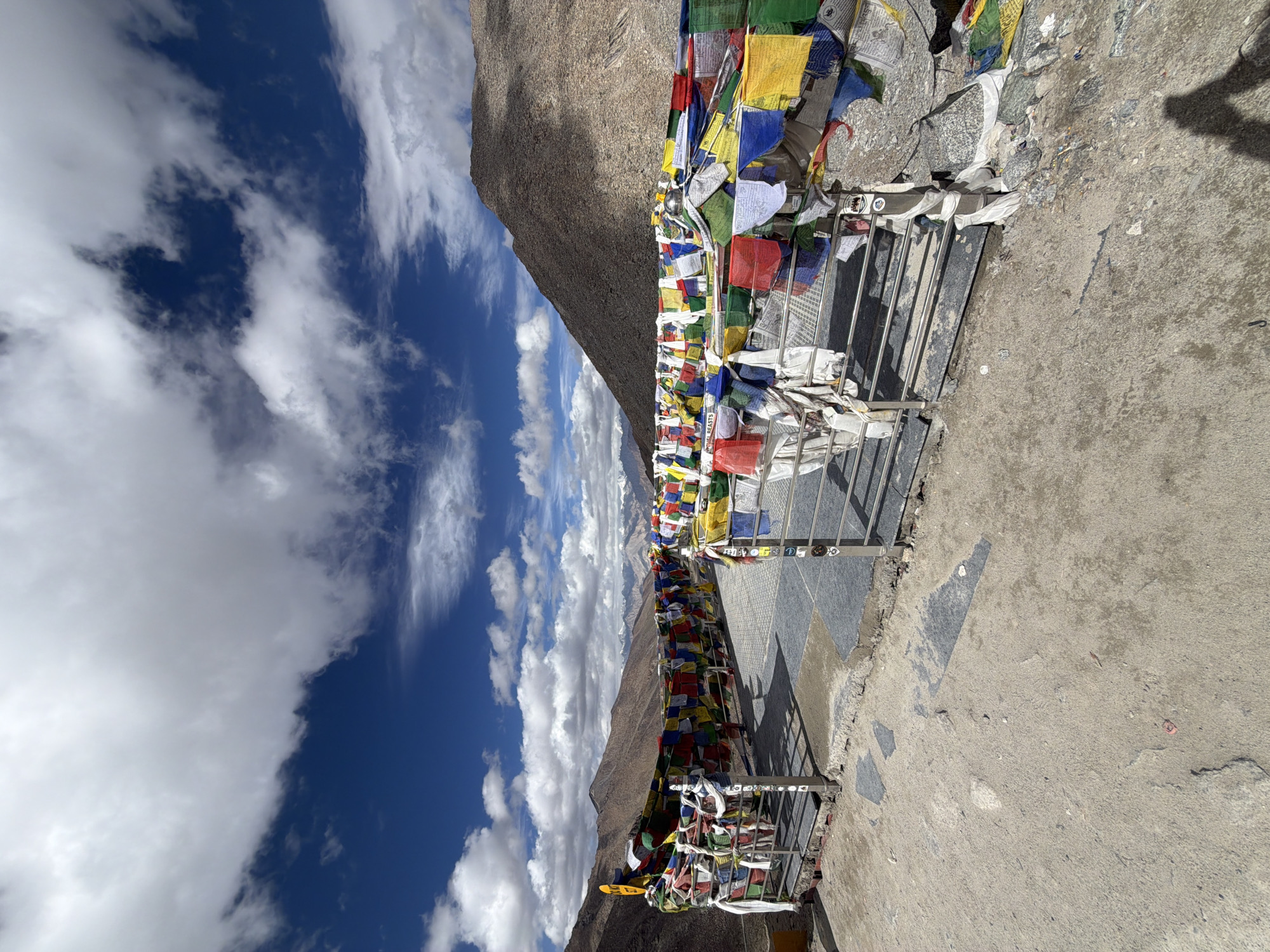 A view from a high vantage point in Leh, India, looking out over a valley. The foreground is a concrete and gravel surface with a metal railing that is extensively decorated with colorful Tibetan prayer flags. These flags, in vibrant reds, blues, yellows, greens, and whites, are tied to the railing and strung across the viewpoint, fluttering in the wind. Behind the railing, a paved walkway extends, also adorned with prayer flags. The mid-ground features a steep, barren, rocky mountain slope on the right, and a less steep, rocky slope on the left. The background reveals a range of majestic, snow-capped mountains under a dramatic sky filled with large, white, cumulus clouds against a deep blue sky. The lighting suggests it is daytime, likely mid-morning or afternoon, casting distinct shadows on the ground. There are no people visible in the frame.