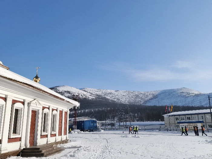 The image depicts an outdoor, snow-covered scene in the city of Im. Gastello, Russia, under a clear blue sky. In the immediate foreground on the left, a portion of a building, likely a church, is visible, featuring white walls with red trim around windows and a visible doorway with steps. Its roof is heavily covered in snow, and a golden onion dome topped with an Orthodox cross rises from the rooftop.
The midground consists of a large, flat expanse of snow with multiple footprints. Several individuals are present: two figures in high-visibility yellow-green jackets and dark pants are seen on the left-mid side, with one appearing to interact with a small white object on the ground. A group of approximately five additional figures, similarly dressed in high-visibility yellow-green jackets and dark pants, walk towards the right on the far right-mid side.
Further in the midground, a large blue container or structure is situated to the left of the walking individuals. Electrical power poles and overhead lines are also visible. In the background on the right, a two-story building with white walls, dark windows, and a dark roof is present, fronted by a low white fence and several flags (red, yellow, blue, white). The distant background features snow-covered mountains with sparse tree cover under a bright, cloudless blue sky, indicating a cold, clear winter day.