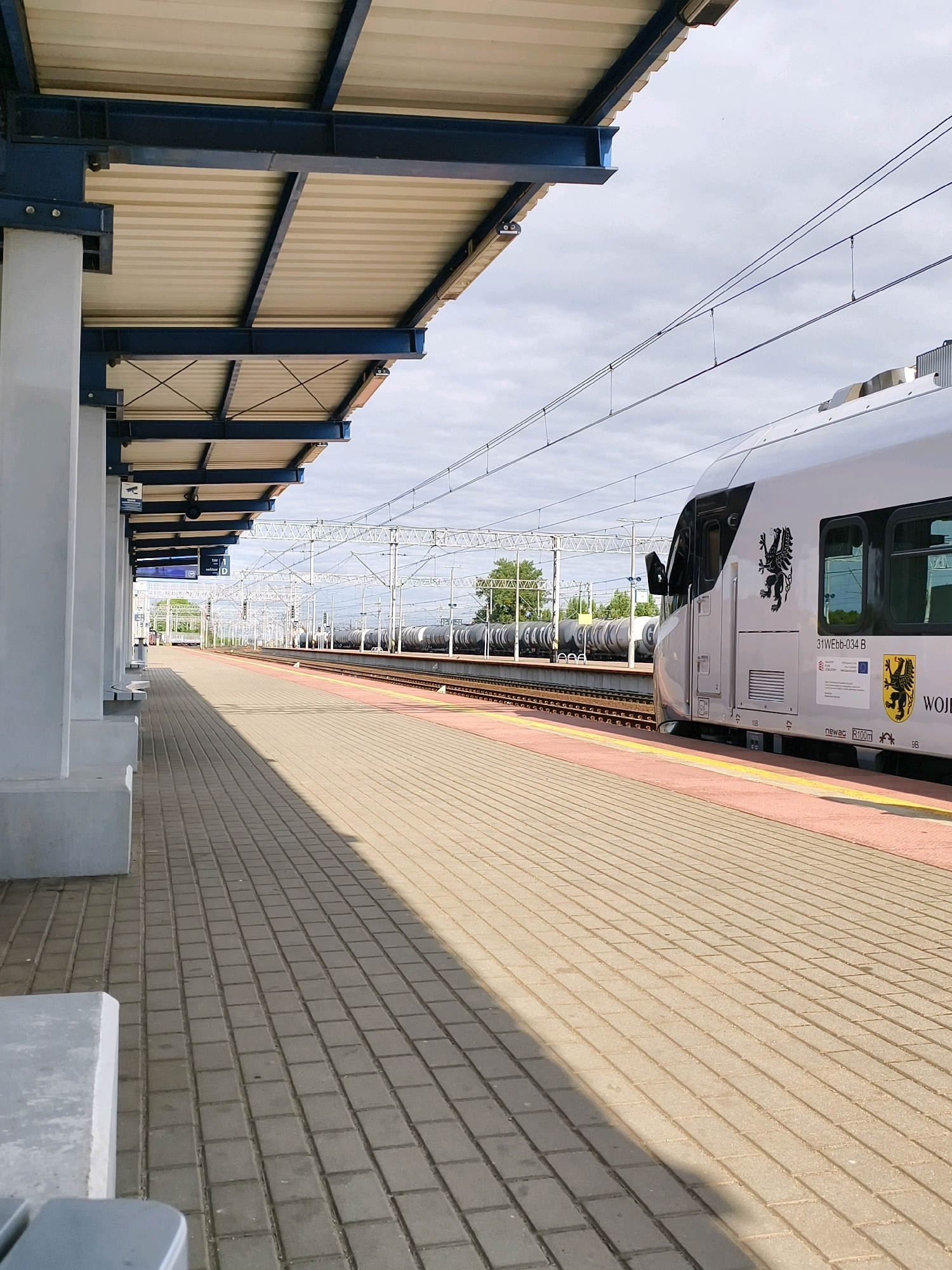 A train is parked at a station platform in Tczew, Poland. The train is white with black accents and features a coat of arms logo. The platform is paved with brick tiles and bordered by a red line. A covered walkway with blue metal beams and a beige roof extends along the platform. Overhead wires are visible above the train tracks. In the background, more trains, trees, and a cloudy sky can be seen. The image appears to be taken from a low angle on the platform, looking down the track.