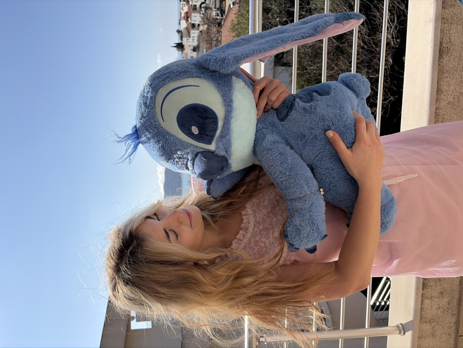 A female with long, wavy blonde hair is depicted outdoors on a balcony or terrace in Budva, Montenegro, under a clear blue sky. She wears a light pink sleeveless top with a textured or lace-like pattern on the upper bodice and a plain light pink lower garment. A silver bracelet with two small white beads is visible on her left wrist. She is holding a large, blue plush toy resembling Disney's Stitch character, which features prominent dark blue eyes, a light blue facial area, a small dark blue nose, and long blue ears with pink inner linings. A small tuft of blue hair protrudes from the toy's head. The female is looking down at the plush toy with a soft smile and her eyes partially closed, cradling it with both arms. In the background, a coastal town featuring white and light-colored buildings with red tiled roofs is visible, interspersed with green vegetation. A body of water, likely the Adriatic Sea, stretches behind the town, with distant hills or mountains on the horizon. A silver-colored metal railing with multiple horizontal bars is in the immediate foreground.