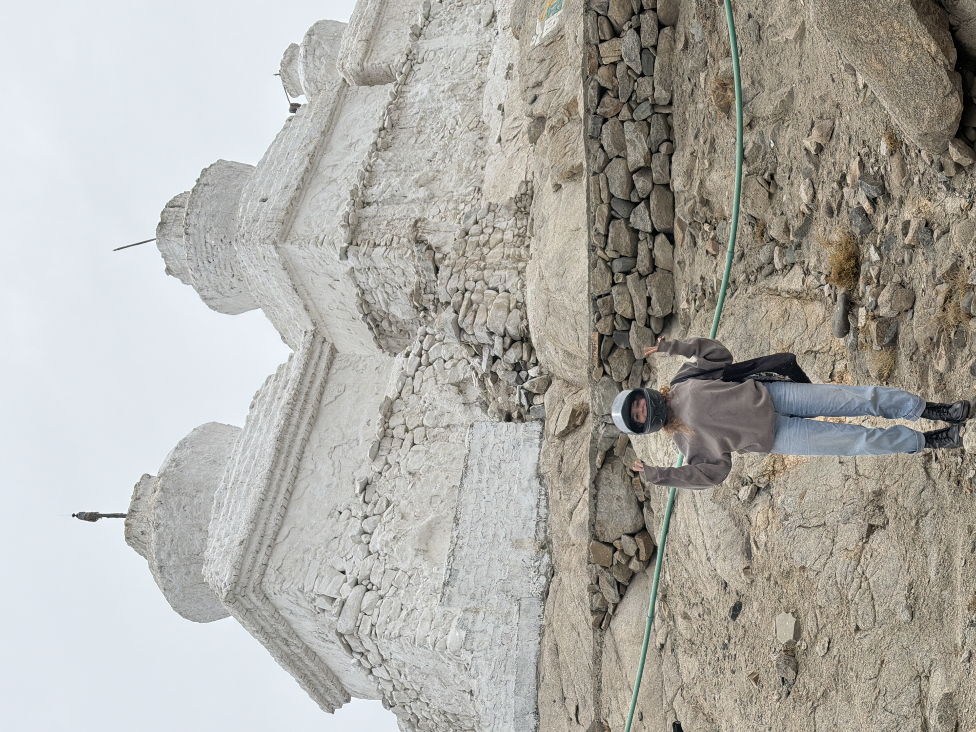 The image shows a young woman standing on a rocky hillside in front of two large, whitewashed stupas. The stupas are tiered structures with rounded tops and appear to be ancient. The woman is wearing a gray sweatshirt, blue jeans, black boots, and a motorcycle helmet. She is holding her hands up in a peace sign. The hillside is covered in rocks and sparse vegetation. The sky is overcast. The location is identified as Thiksey, India, which is known for its large monastery with similar architectural features.
