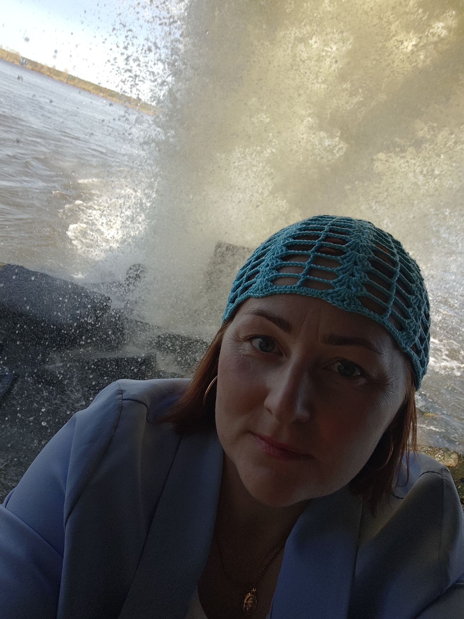 A woman is taking a selfie in what appears to be an outdoor setting with a body of water in the background. The woman has fair skin, reddish-brown hair, and is wearing a blue crocheted cap. She is also wearing a blue jacket, a gold necklace with a medallion, and hoop earrings. The background shows a body of water with waves crashing against what could be rocks or a barrier. Splashes of water are visible, suggesting a potentially windy or turbulent environment. Based on visual clues, it is impossible to confirm the location as Chernushka, Russia, or any specific location.