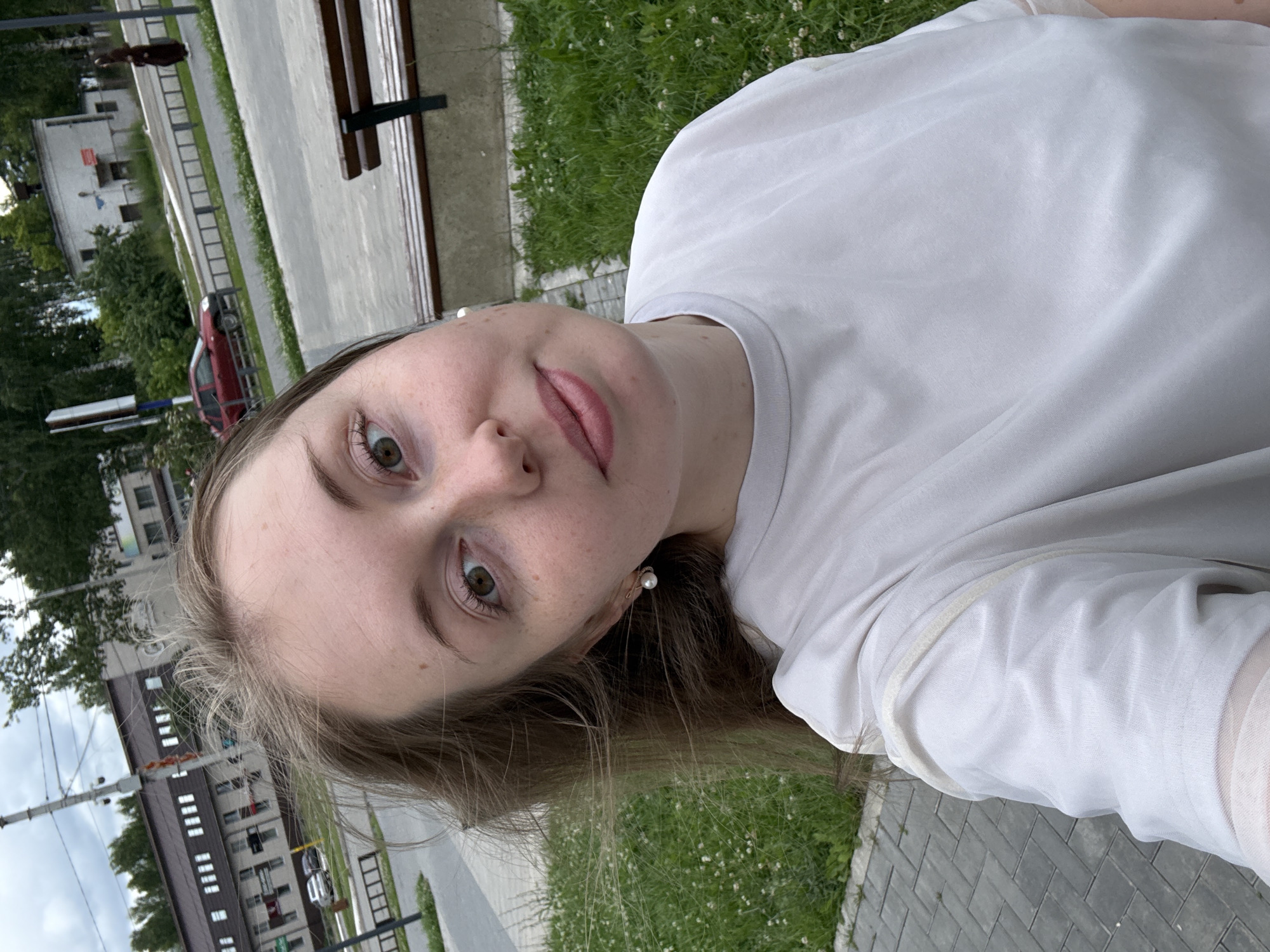 A young woman with light brown hair and fair skin takes a selfie in an urban park. She is wearing a light grey t-shirt and a pearl earring in her left ear. Her face is turned towards the camera, and she is looking directly at it. Behind her, a paved walkway is visible, as well as a grassy area with small white flowers. In the background, there are buildings, trees, a red car, and a park bench. The sky is overcast.