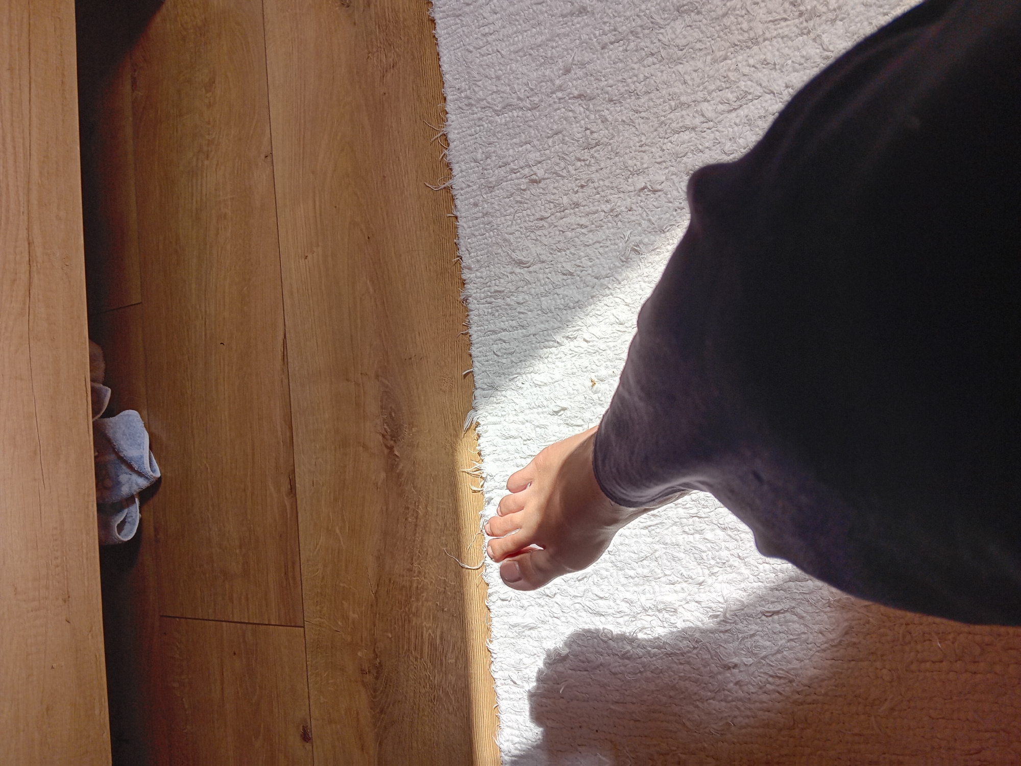 An overhead shot shows a bare foot and lower leg resting on a white, textured rug. The foot is positioned at an angle, with the toes pointing towards the upper left of the frame. Sunlight streams in from the left, casting a shadow of the foot and leg onto the rug. Behind the rug, a wooden floor is visible, with a darker wooden surface at the top of the frame, possibly a piece of furniture. Underneath this darker surface, a small, light blue towel is partially visible. The scene appears to be indoors, likely in a residential setting, and no specific landmarks or features indicating a location in Zuberec, Slovakia, are discernible.
