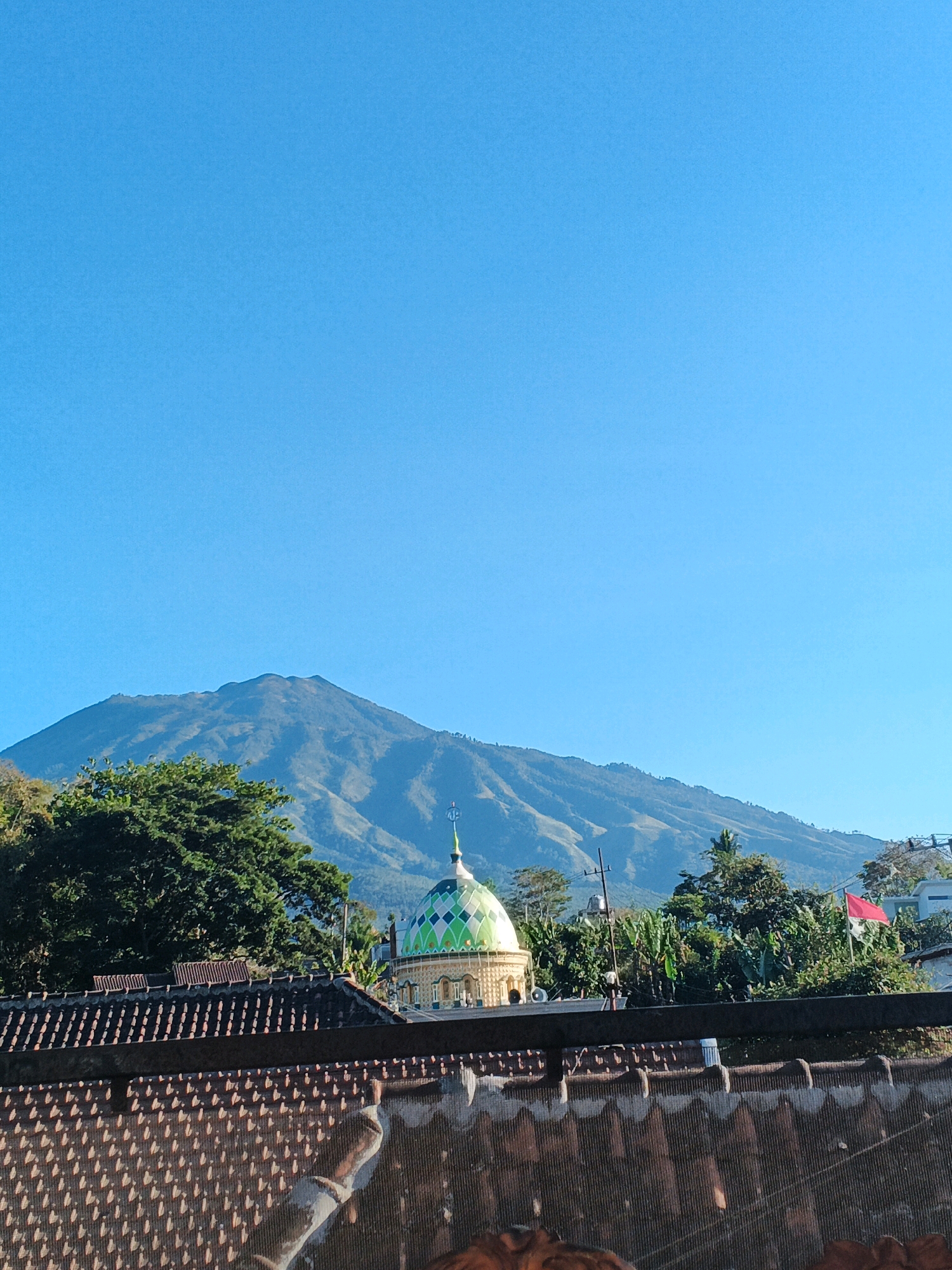 An image depicts a landscape view with a prominent mountain range in the background under a clear blue sky. In the foreground, several elements are visible, including a tiled roof, trees, and a mosque with a green and white checkered dome. A red flag is also seen flying from a pole amidst the greenery. The scene appears to be a village setting, possibly in Bocek, Indonesia, given the architectural style and the presence of lush vegetation. The lighting suggests it is daytime, with the sun casting shadows and illuminating the landscape. No people are visible in the image.
