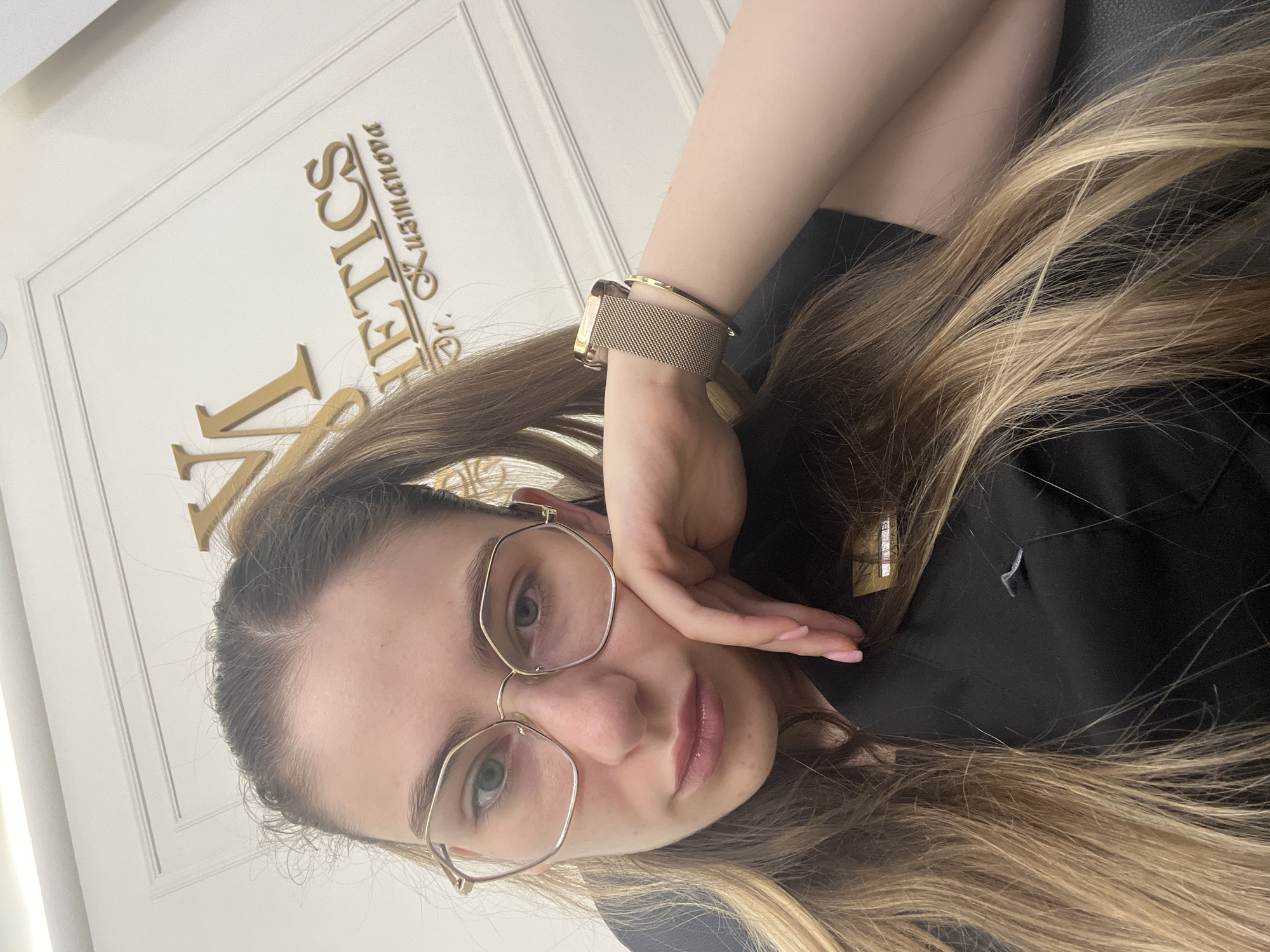 A young Caucasian woman with long, blonde hair pulled into a ponytail is seen in a close-up, slightly angled selfie. She is wearing large, gold-rimmed, hexagonal glasses and a black scrub top. Her left arm is propped up, with her hand resting near her chin, and she is wearing a gold mesh Apple Watch band and a gold bangle. Behind her, on a white, paneled wall, is a gold embossed sign with stylized letters that reads "VM KUZMANOVA." The lighting appears to be artificial and bright, suggesting an indoor setting. The woman's expression is neutral.