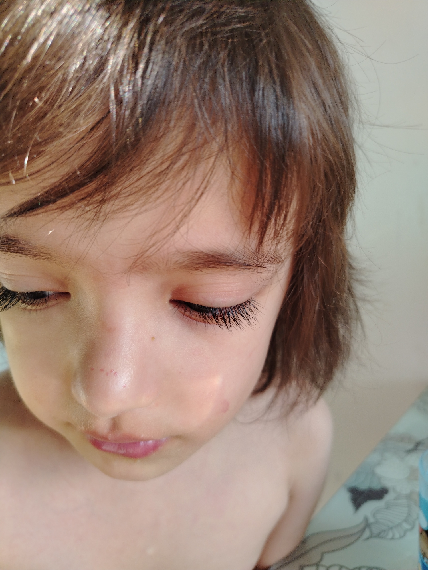 A close-up shot of a young child with fair skin and light brown hair. The child's eyes are closed or cast downwards, displaying long, dark eyelashes. The face is illuminated by a soft, natural light. The background is blurred and indistinct but appears to be a neutral-colored wall. No specific location in Noviy, Russia is discernible from the image.