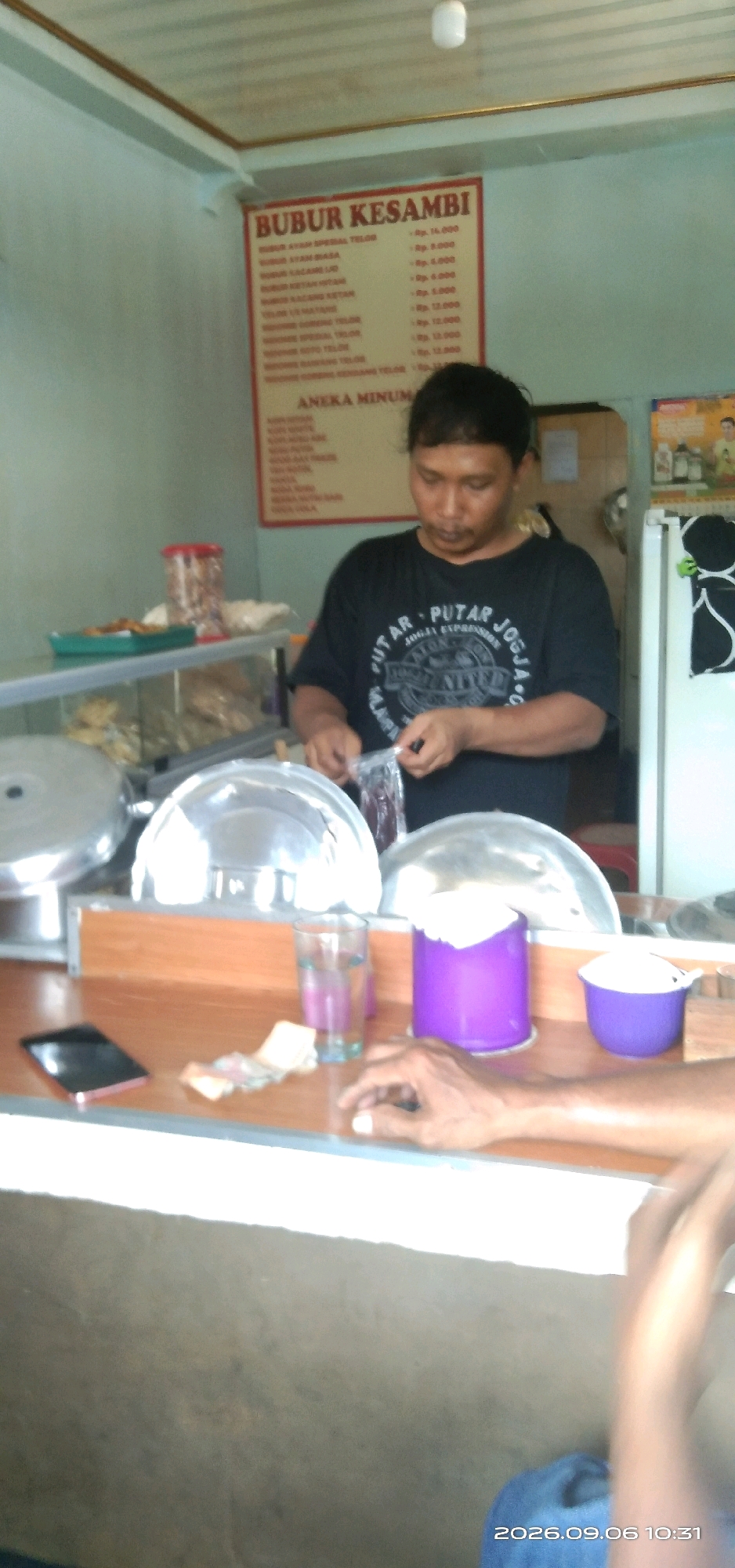A man stands behind a counter in what appears to be a small eatery in Cirebon, Indonesia. He is wearing a black t-shirt with a graphic design and is in the process of opening a plastic package. In front of him on the counter are several metallic bowls, a glass of purple liquid, a purple container, and a small purple bowl. There is also a smartphone and some Indonesian currency on the counter. A menu board is visible on the wall behind him, listing various food and drink items. The environment suggests a casual, local establishment.