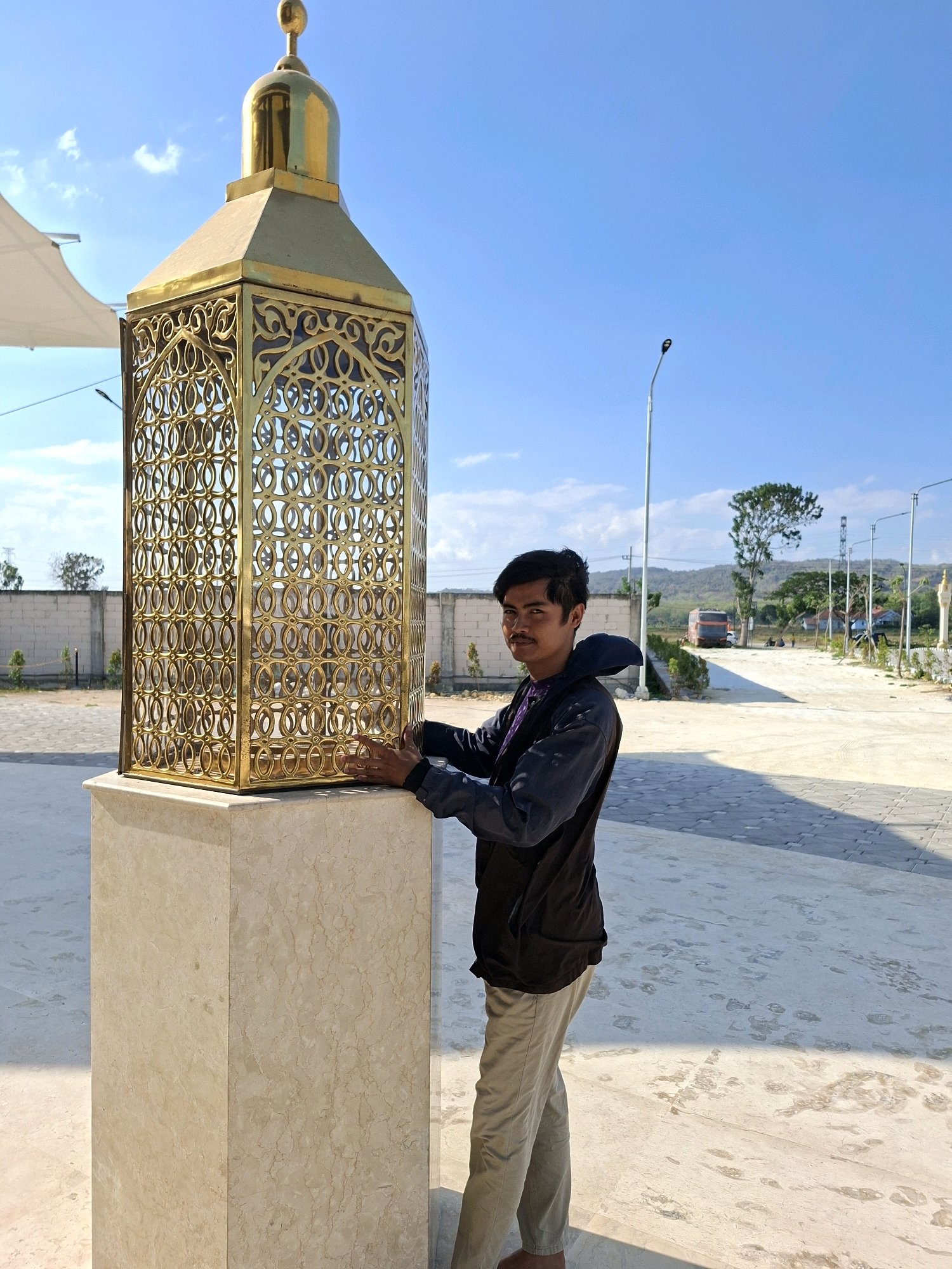 A young man stands next to a large, ornate golden lantern in Jrengik, Indonesia. The lantern is octagonal in shape with intricate geometric carvings and a pointed golden dome. It sits on a light-colored marble pedestal. The man is wearing a dark jacket over a purple shirt and light-colored pants. His hands are resting on the lantern. The background shows a paved outdoor area with a white brick wall and some trees. In the distance, a bus, streetlights, and a hazy horizon with hills are visible under a bright blue sky.