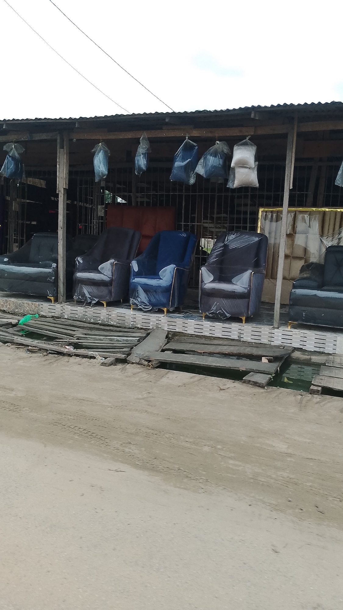The image shows a display of furniture outside a shop in an urban environment. There are five upholstered chairs visible, arranged in a row on an elevated platform. The chairs are covered in clear plastic wrapping. The chair fabrics appear to be blue and dark gray. Behind the chairs, the shop has a metal grill facade, and some items are hanging from the roof. In front of the chairs, there is a makeshift wooden walkway leading to the street. The street is unpaved and appears sandy. The location is an unknown city, in an unknown state/province.
