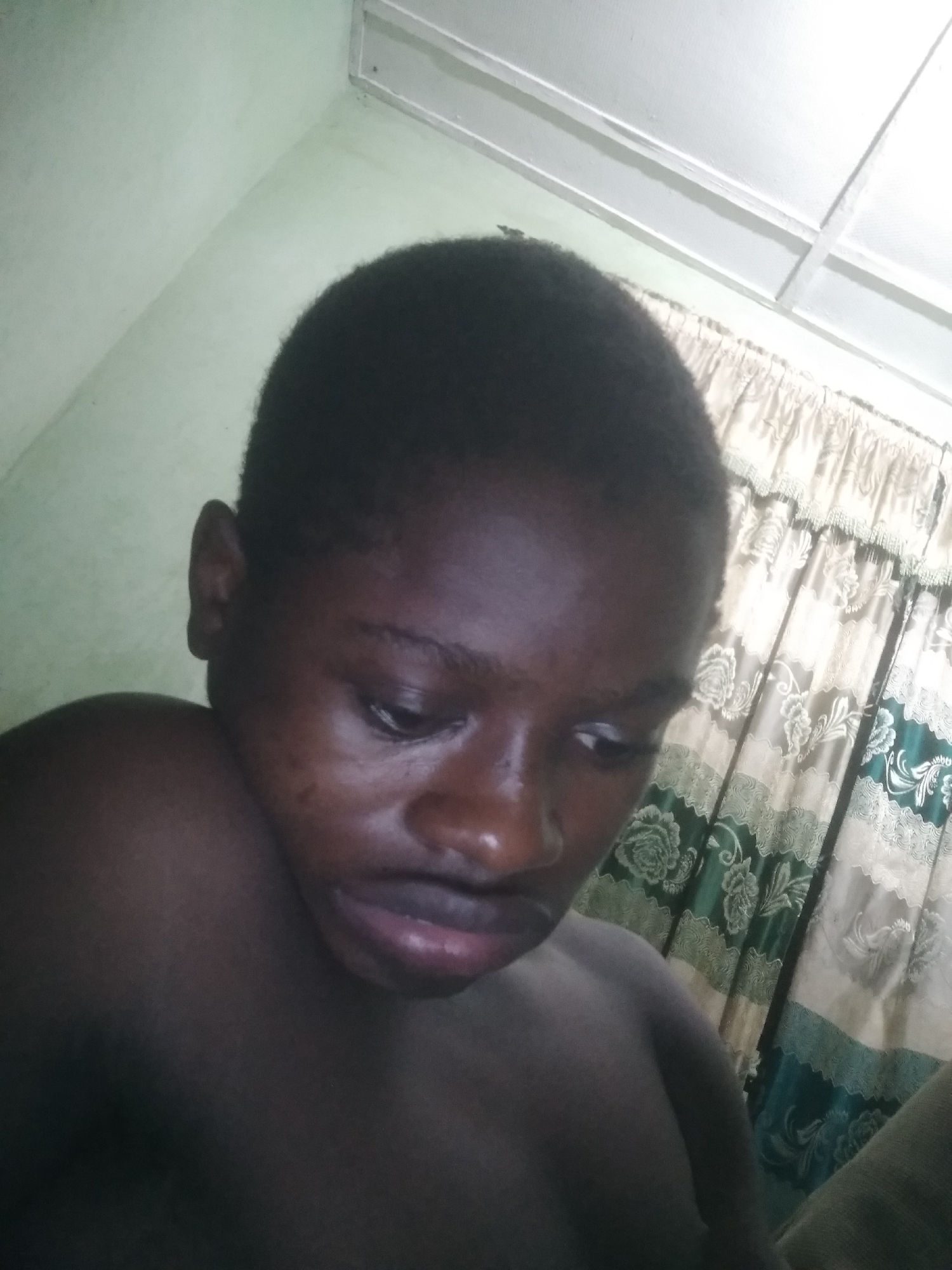 The image shows a dark-skinned person with a closely cropped haircut, looking downward. The person appears to be shirtless. The background includes a light green wall and a window with patterned curtains. The location is not evident in the image, and cannot be verified to be in the city of Shangisha, Nigeria.