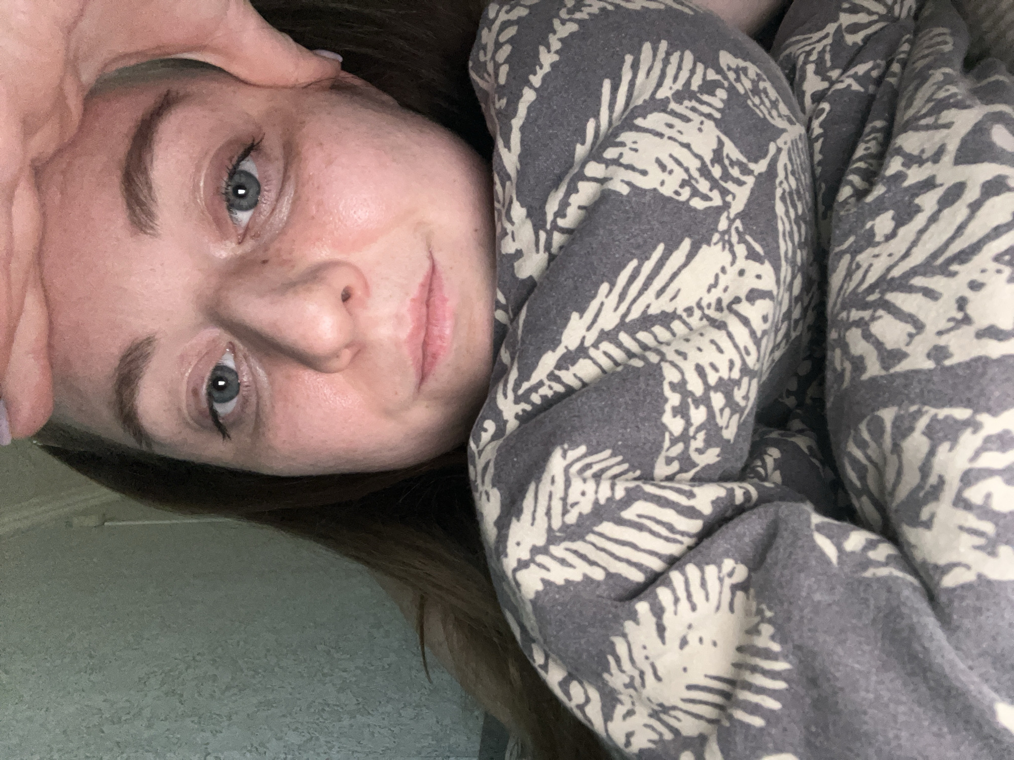 A woman with fair skin, light blue eyes, and brown hair is close to the camera, with part of her face obscured by her hand. Her eyebrows are slightly raised. She is partially covered by a gray blanket with a white leafy pattern. The background appears to be a simple light-colored wall. It is unclear how the image is related to the city of Раздолье, Russia.