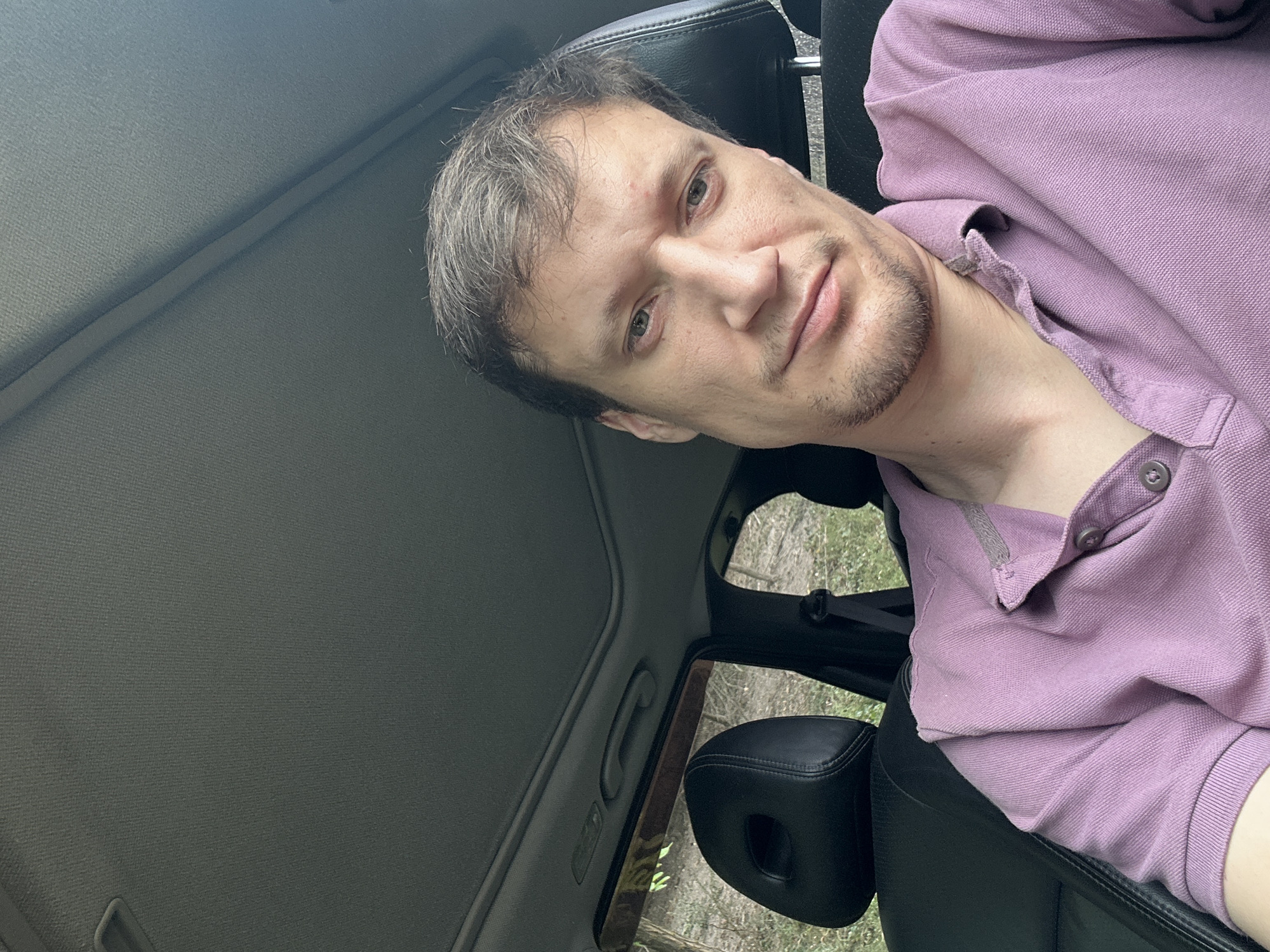 A man is sitting in the driver's seat of a car. The man has light brown hair, light skin, and a light brown beard. He is wearing a purple polo shirt. The car has a sunroof that is open. The car is parked on a dirt road with trees and grass visible outside the windows.
