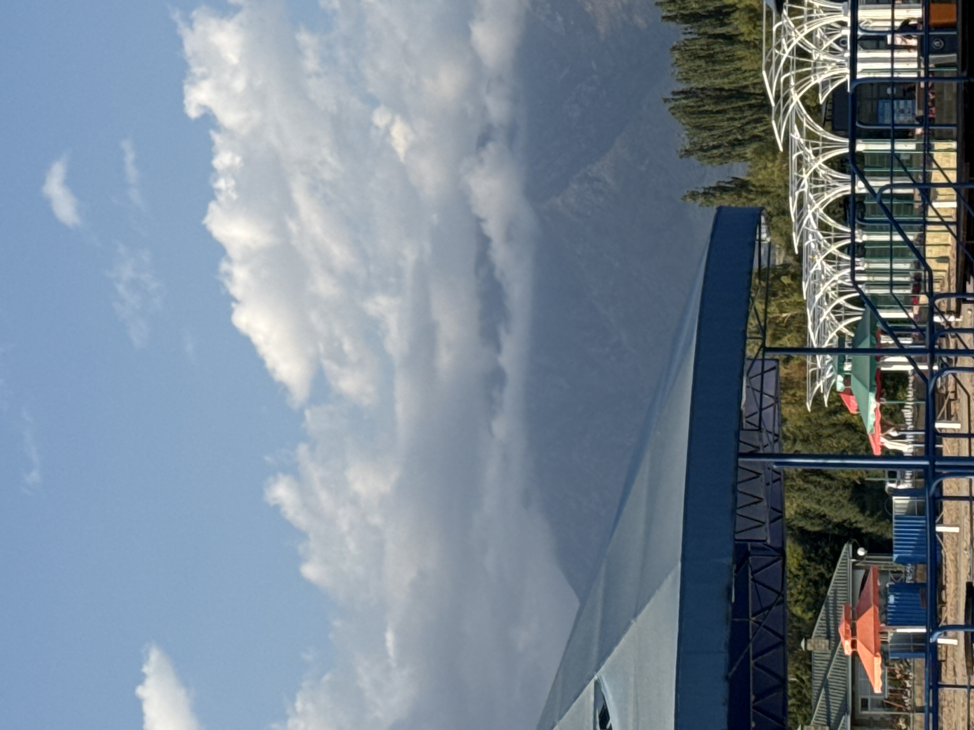 In Sary-Oy, Kyrgyzstan, a scene unfolds with a focus on an outdoor recreational area. In the foreground, a large blue canopy structure dominates, offering shade. Below this, a row of white arched structures with translucent roofing stretches across the mid-ground. To the left, an orange umbrella is visible over a building with a peaked roof. A stand of tall, slender trees lines the horizon behind the arched structures. In the distance, imposing mountains rise, partially obscured by a layer of soft, white clouds. The sky above is a clear blue, dotted with more scattered white clouds. The overall impression is one of a sunny day at a resort or park.