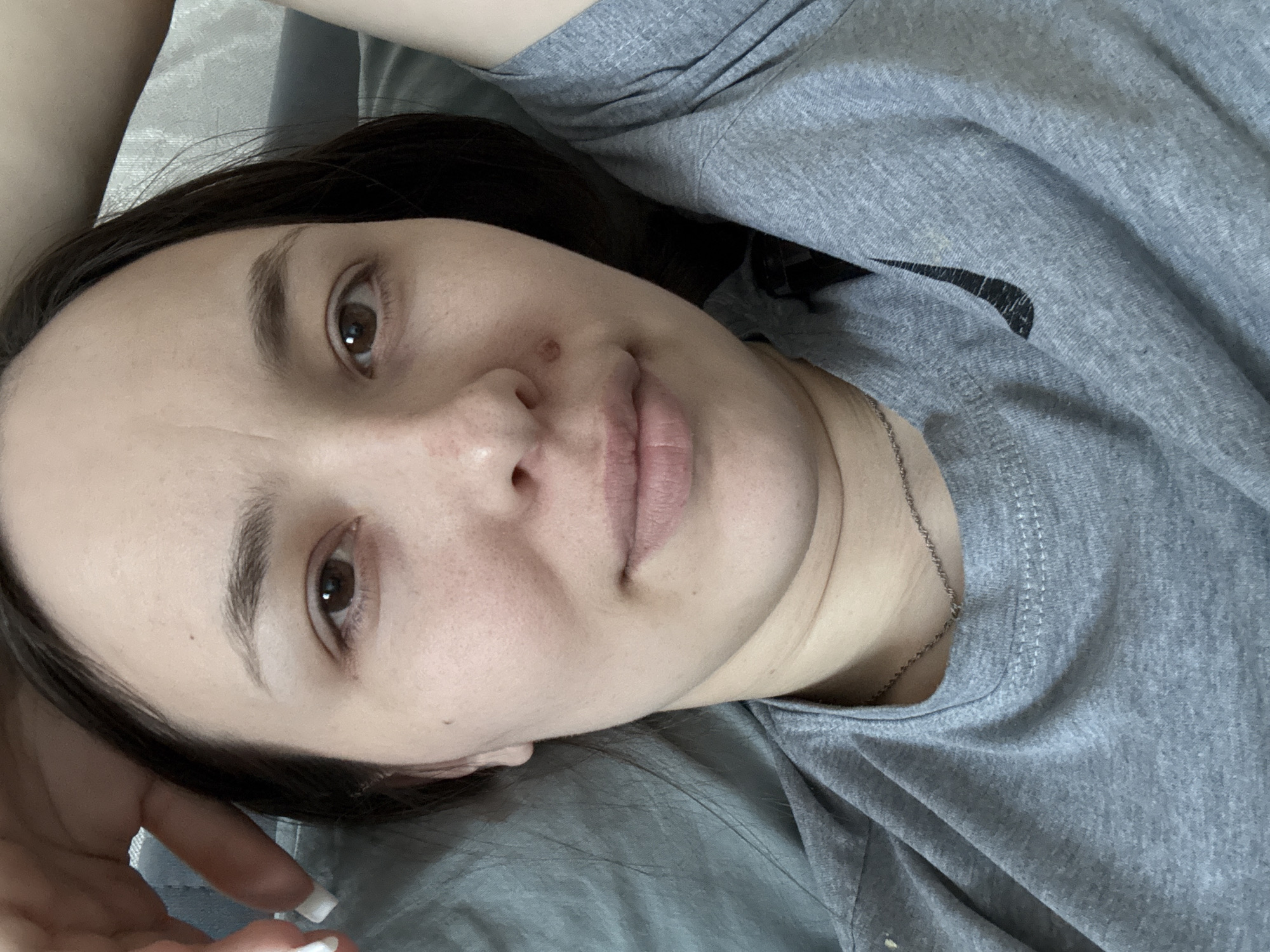 A woman is lying on a gray surface, likely a bed, wearing a gray T-shirt with a black Nike swoosh visible on the right side of the frame. She has dark hair, brown eyes, and light skin. A small mole is visible on her face near her nose. She is wearing a thin silver necklace. Her left hand is slightly visible in the lower left corner of the frame with white nail polish. There is no information to determine a location in the city of Chaikovsky, Russia.
