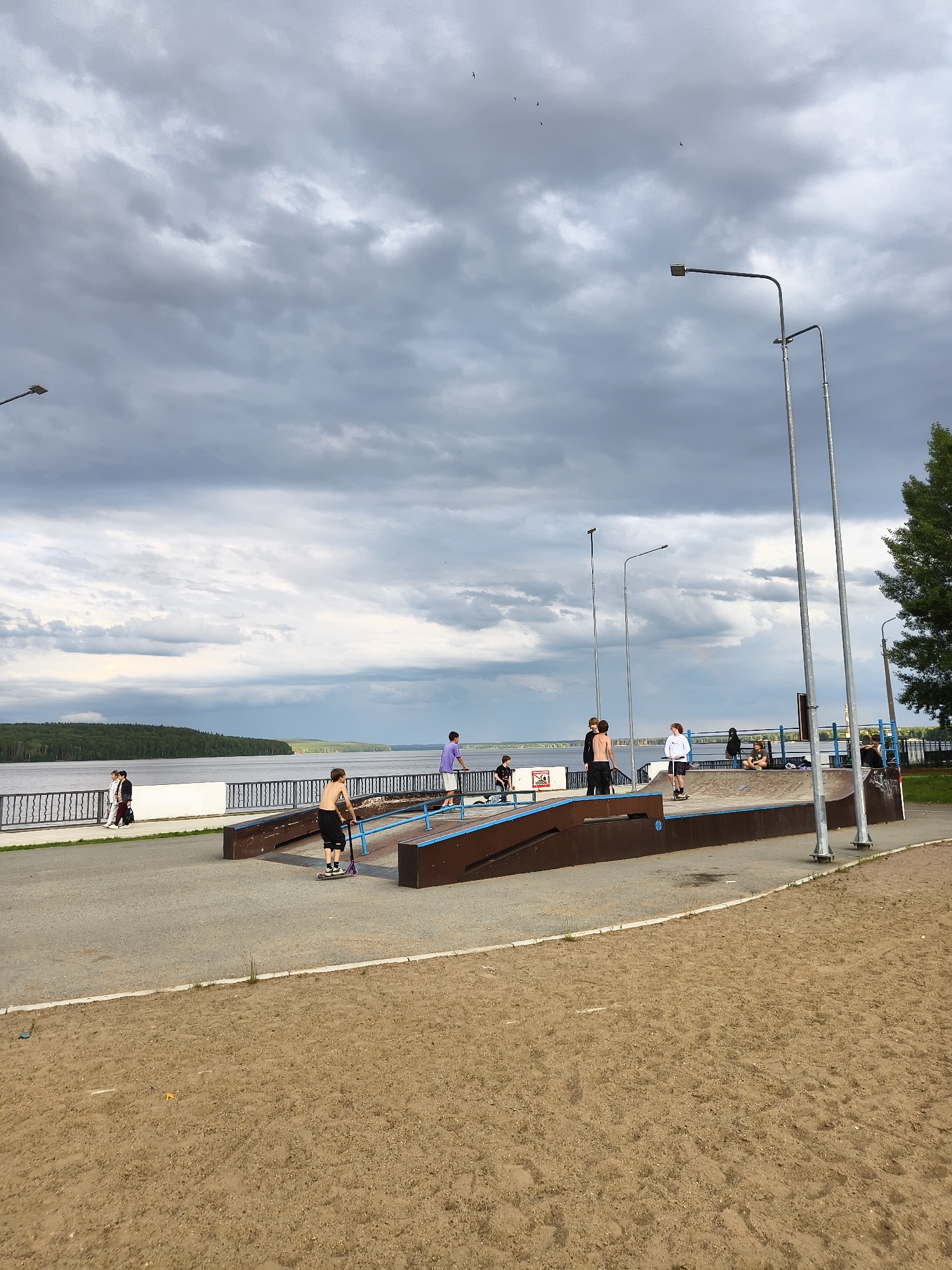 The image depicts a skatepark on a cloudy day, situated near a large body of water. The sky is filled with dramatic, gray clouds, suggesting an overcast or potentially stormy atmosphere.
Several people are present at the skatepark, engaging in various activities. A young person is seen on a scooter, positioned on a ramp. Other individuals are scattered around the skatepark, some observing, others appearing to rest or converse. The skatepark itself features a series of ramps and transitions, characteristic of a facility designed for skateboarding and similar sports.
In the background, a wide river or lake stretches across the frame, with a forested shoreline visible on the opposite bank. A railing separates the skatepark area from the waterfront. Two streetlights stand tall near the skatepark. The ground in the foreground is a sandy area, typical of an outdoor recreational space.
The overall mood appears casual and relaxed, despite the ominous sky. The lighting suggests it is daytime, likely late afternoon or early evening given the cloud cover. The presence of the river and the architecture hints at a potentially a public park or waterfront recreational area in Chaikovsky, Russia. There is no visible text in the image.