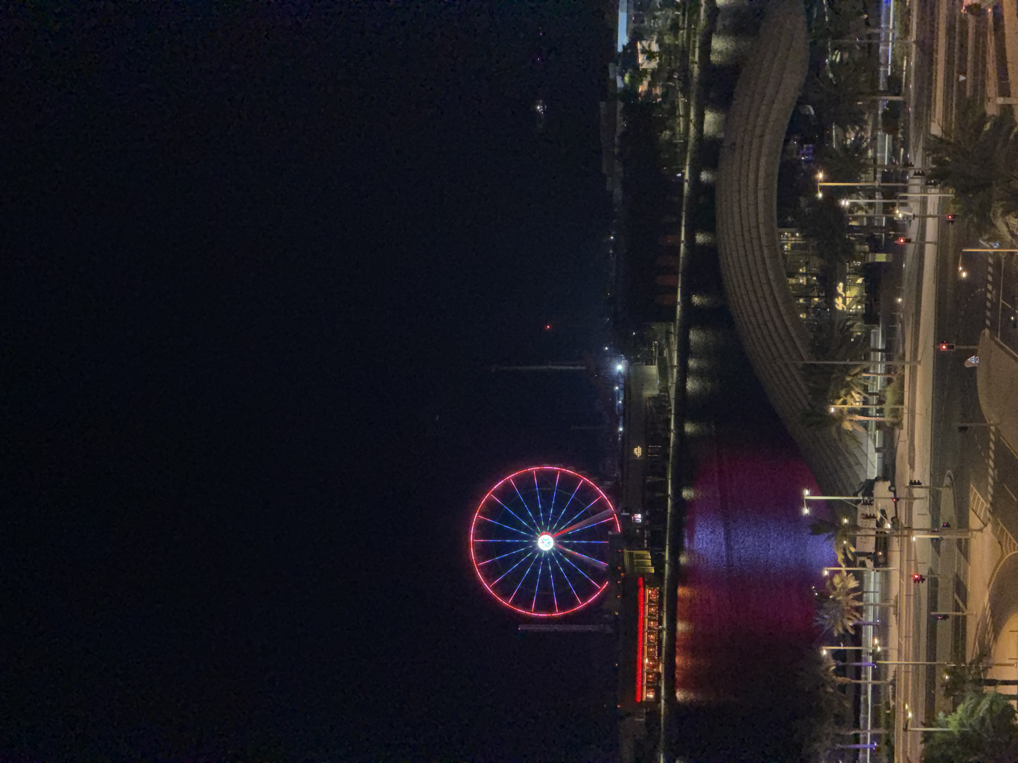 A Ferris wheel illuminated with red and blue lights stands prominently against a dark night sky in the city of Unknown. The wheel, a large circular structure, is supported by a tall, slender tower. In front of the Ferris wheel, a body of water reflects the colorful lights of buildings and the Ferris wheel itself, creating vibrant hues of red and blue on its surface. Along the waterfront, a row of buildings with red illuminated facades and other structures are visible, suggesting a commercial or entertainment district.
Further in the foreground, a modern, curved architectural structure with a patterned roof dominates the scene. Palm trees line the area around this structure, and streetlights cast a warm glow on the surroundings. A road with traffic lights and pedestrian crossings is visible, with a few vehicles, including trucks and vans, present. The overall environment depicts a well-lit urban landscape at night, likely a tourist or recreational area, with the Ferris wheel serving as a central landmark.