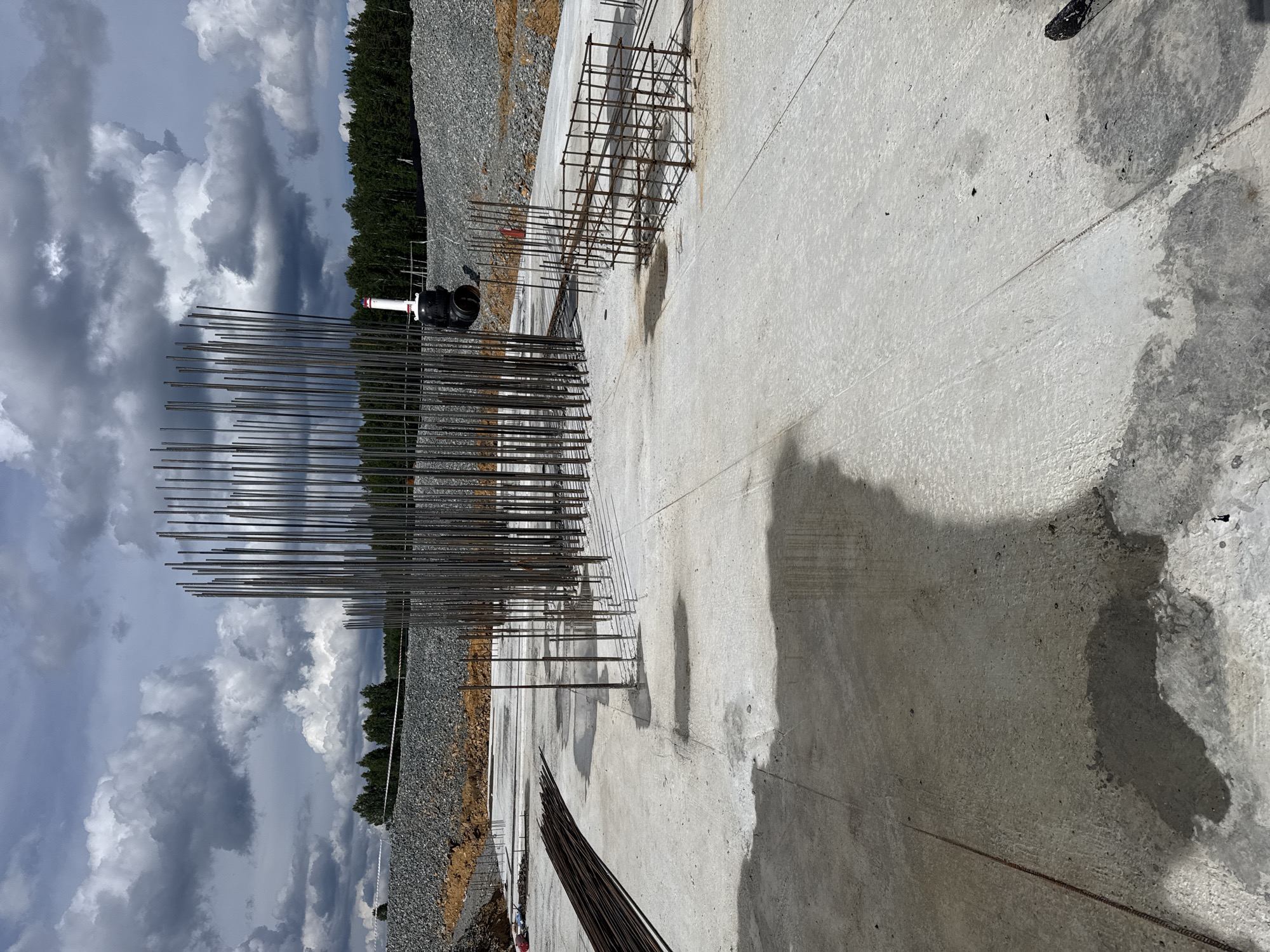 The image captures an outdoor construction scene, likely in Leninsk, Russia, under a dramatic sky filled with heavy, dark clouds interspersed with brighter patches, suggesting a change in weather. The foreground is dominated by a large expanse of wet, grey concrete, showing dark, irregular water stains and some lighter, smoother patches. A prominent feature is a dense arrangement of vertical steel rebar, sticking up from the concrete surface. To the left of this rebar cluster, a black pipe fitting with a white element attached is visible, positioned on the concrete near the edge of the construction site.

Beyond the concrete, a steep slope covered in gravel and dirt leads up to a dense line of dark green trees. The overall impression is one of ongoing construction work in a somewhat overcast environment. The time of day is not definitively clear, but the shadows cast on the concrete suggest it is daytime. The presence of rebar and concrete points to the foundation or structural work of a building or infrastructure project. No text is visible in the image.
